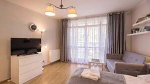 A modern hotel room with a flat-screen TV, a sofa, a bed with a blanket, a small table, and a window with curtains. - Rosa Weneda by Baltic Home (Swinoujscie)