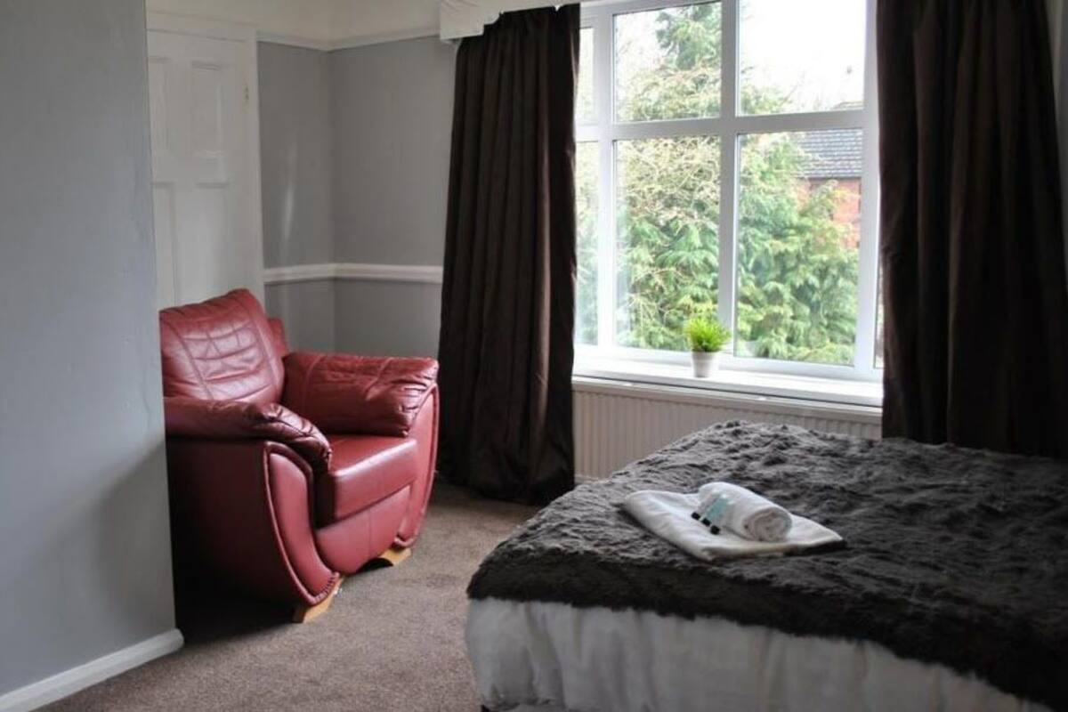 A bedroom with a bed, a red leather chair, a window with curtains, and a small potted plant.
