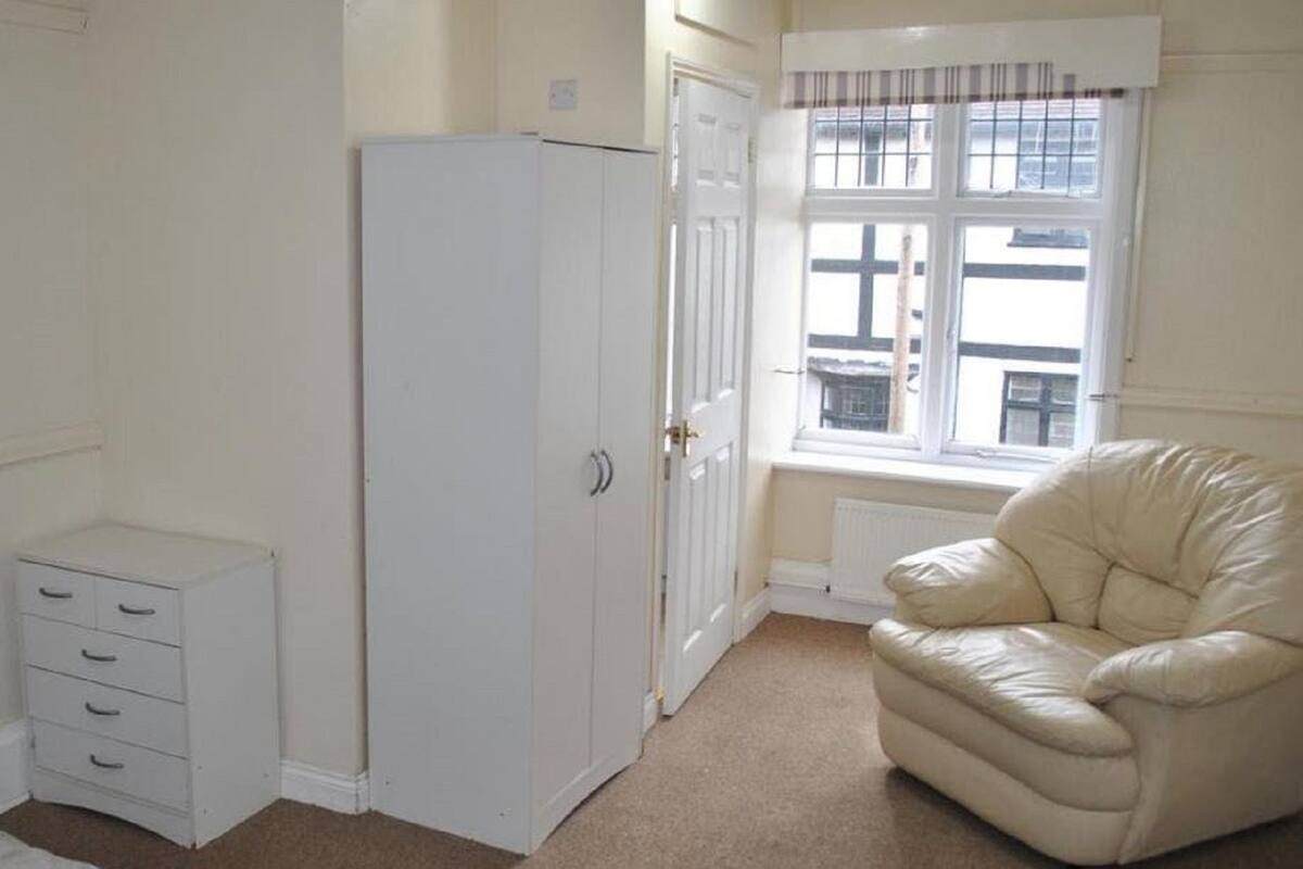 A room with a white wardrobe, a small chest of drawers, a window with blinds, and a leather armchair.