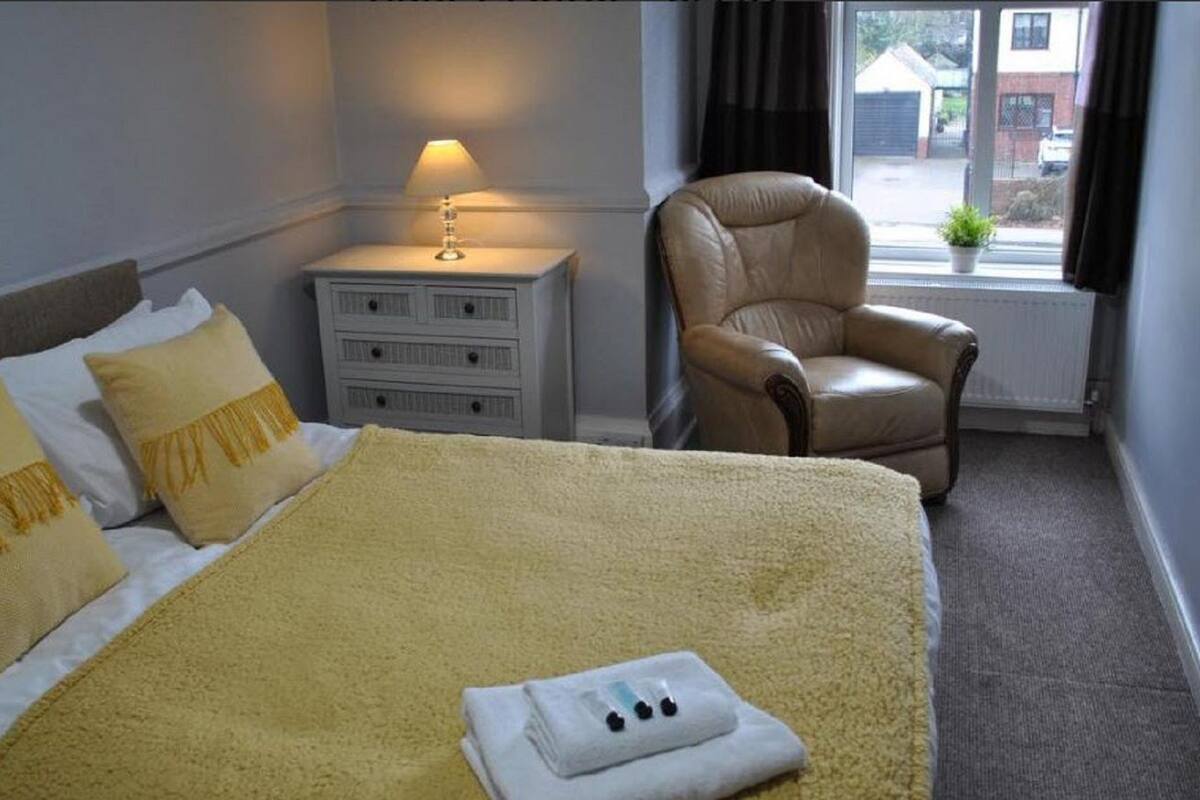 A hotel room with a bed, a chair, a nightstand, a lamp, and a window with a view of the outside.