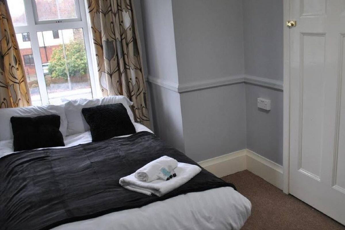 A bedroom with a bed, a window with curtains, a door, and a light switch on the wall.