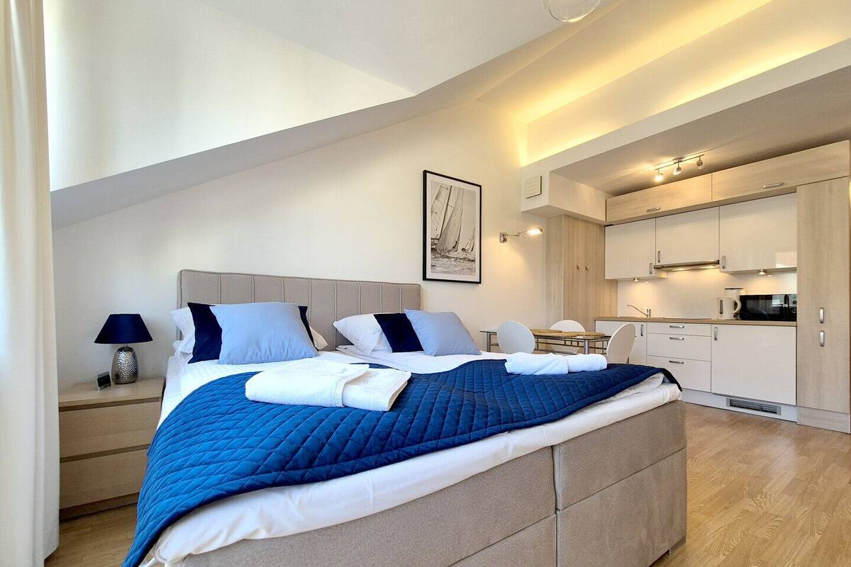 A modern bedroom with a large bed, a bedside table, a lamp, and a view of the kitchen area.