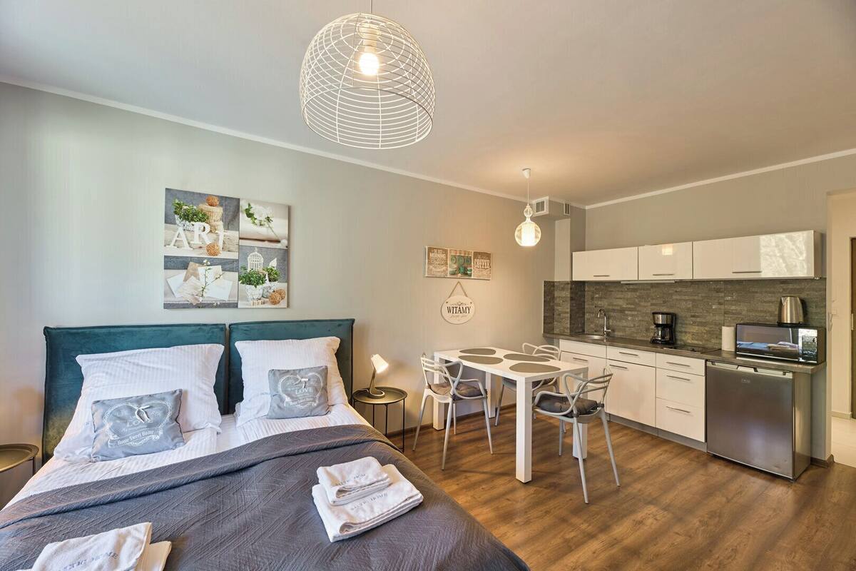 A modern bedroom with a bed, bedside tables, and a small dining table with chairs. The room has a kitchenette with a stove, microwave, and cabinets.