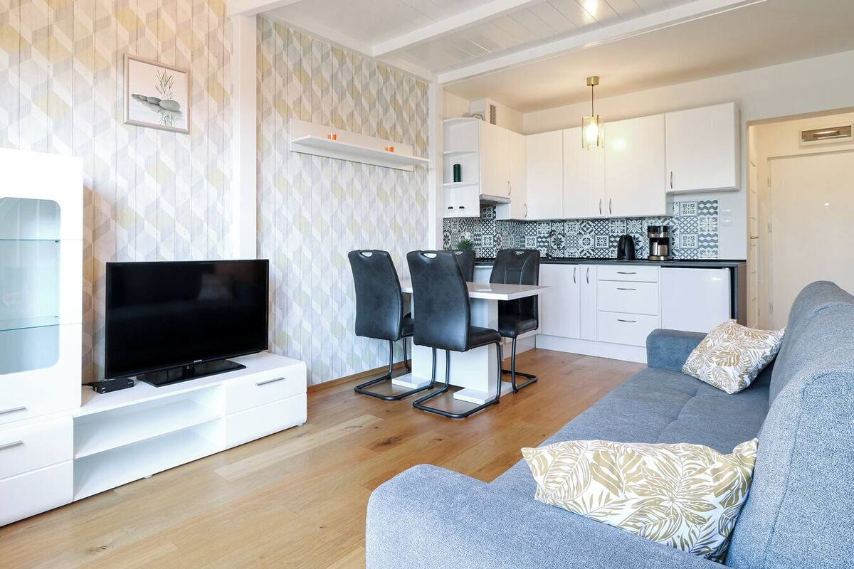 A modern living room with a flat-screen TV, grey sofa, and patterned cushions. A dining area with a table and chairs is adjacent to the kitchen, which features white cabinets and a black and white backsplash.