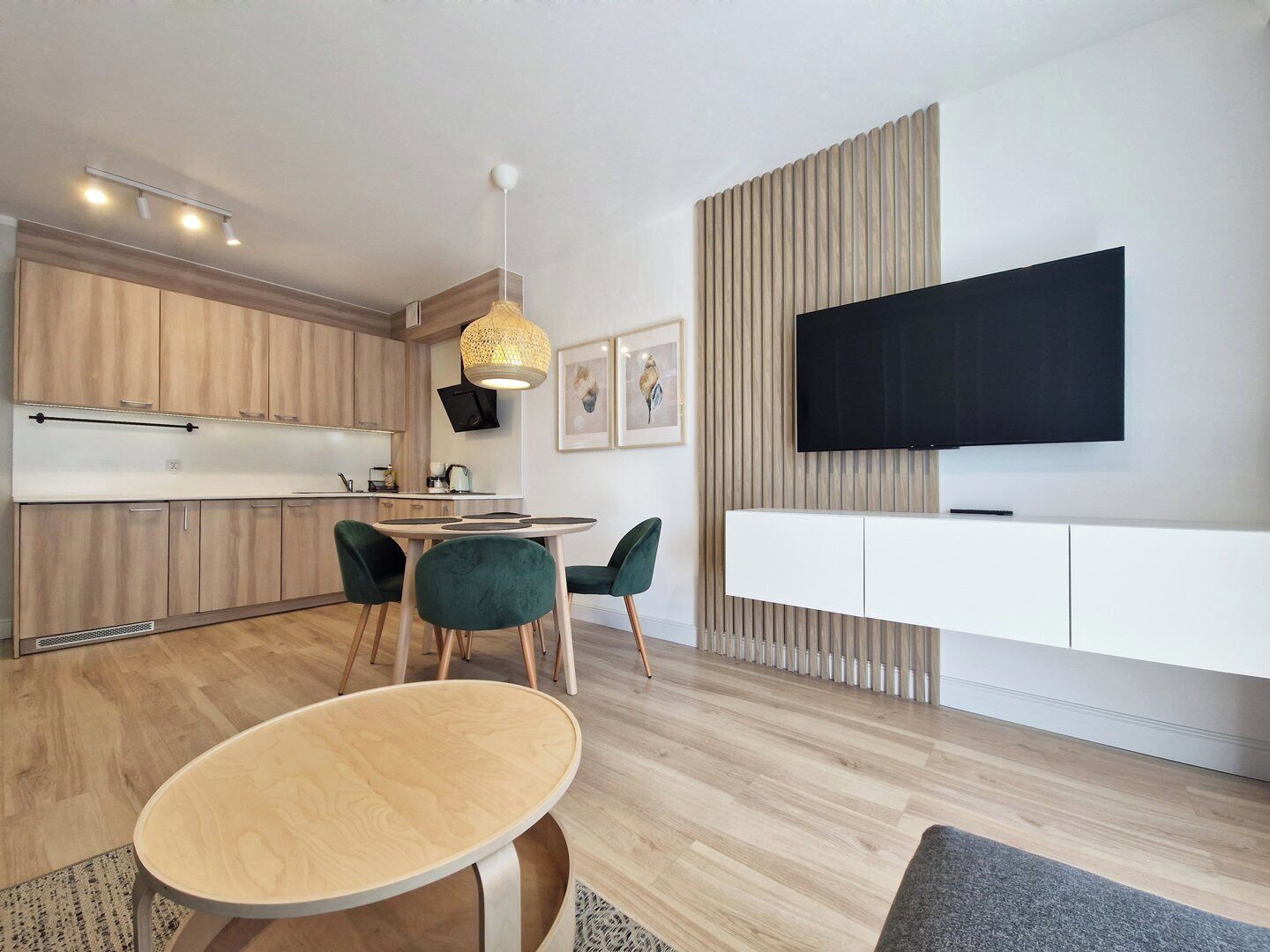 Una cocina moderna con armarios de madera, un comedor con mesa redonda y sillas verdes, un televisor de pantalla plana montado en una pared blanca, y una lámpara en el techo.