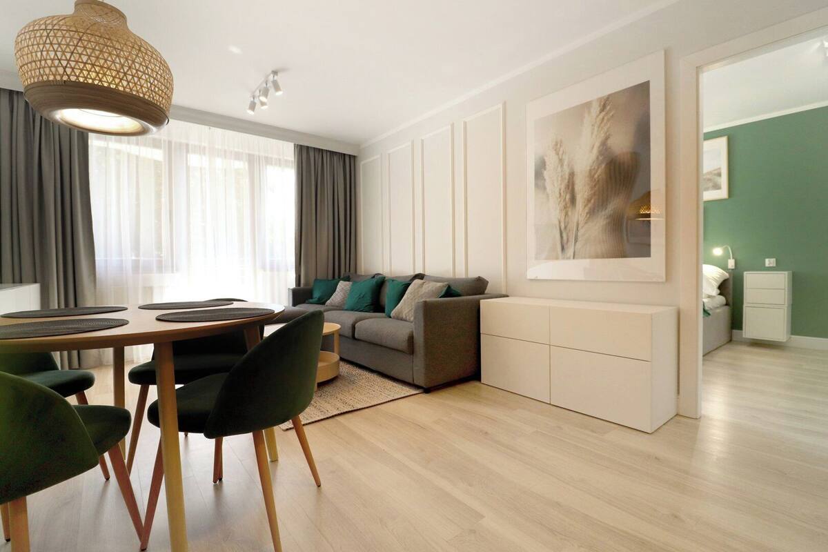 A modern living room with a dining area, a grey sofa with green cushions, a wooden dining table, and a hanging light fixture.