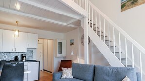 Un vaste espace de vie avec un canapé gris, une cuisine à l’arrière-plan et un escalier blanc.