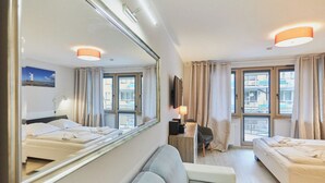Une chambre d’hôtel avec deux lits, un canapé, un bureau, une chaise, un miroir et de grandes fenêtres avec des rideaux.