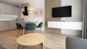 Une cuisine moderne avec des meubles en bois, un coin repas avec une table ronde et des chaises vertes, un téléviseur à écran plat fixé au mur et un canapé gris dans le salon.