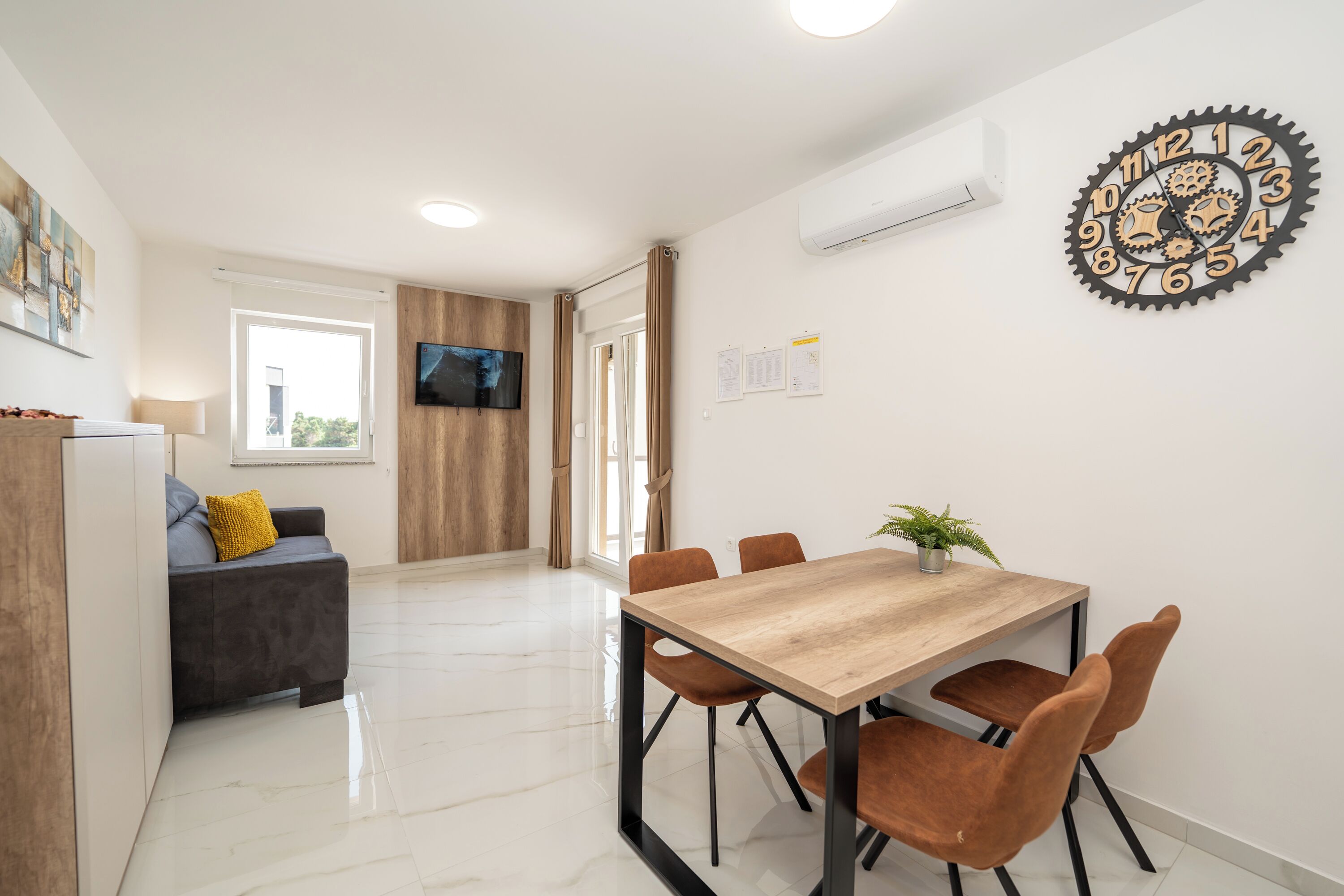 A modern kitchen with a wooden table and chairs, a TV mounted on the wall, and a window with curtains.