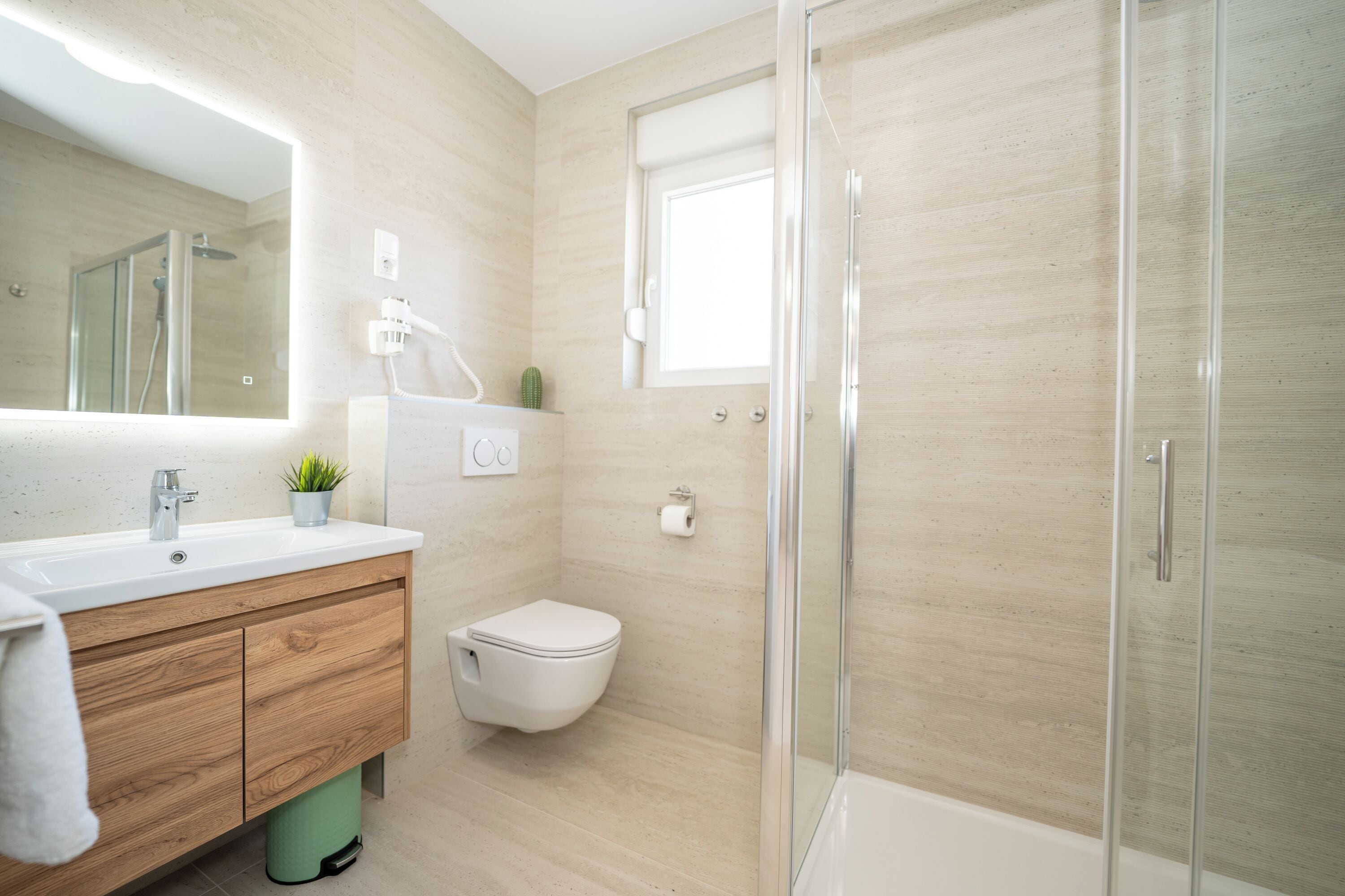 A modern bathroom with a wooden vanity, a toilet, a glass shower enclosure, and a small window.