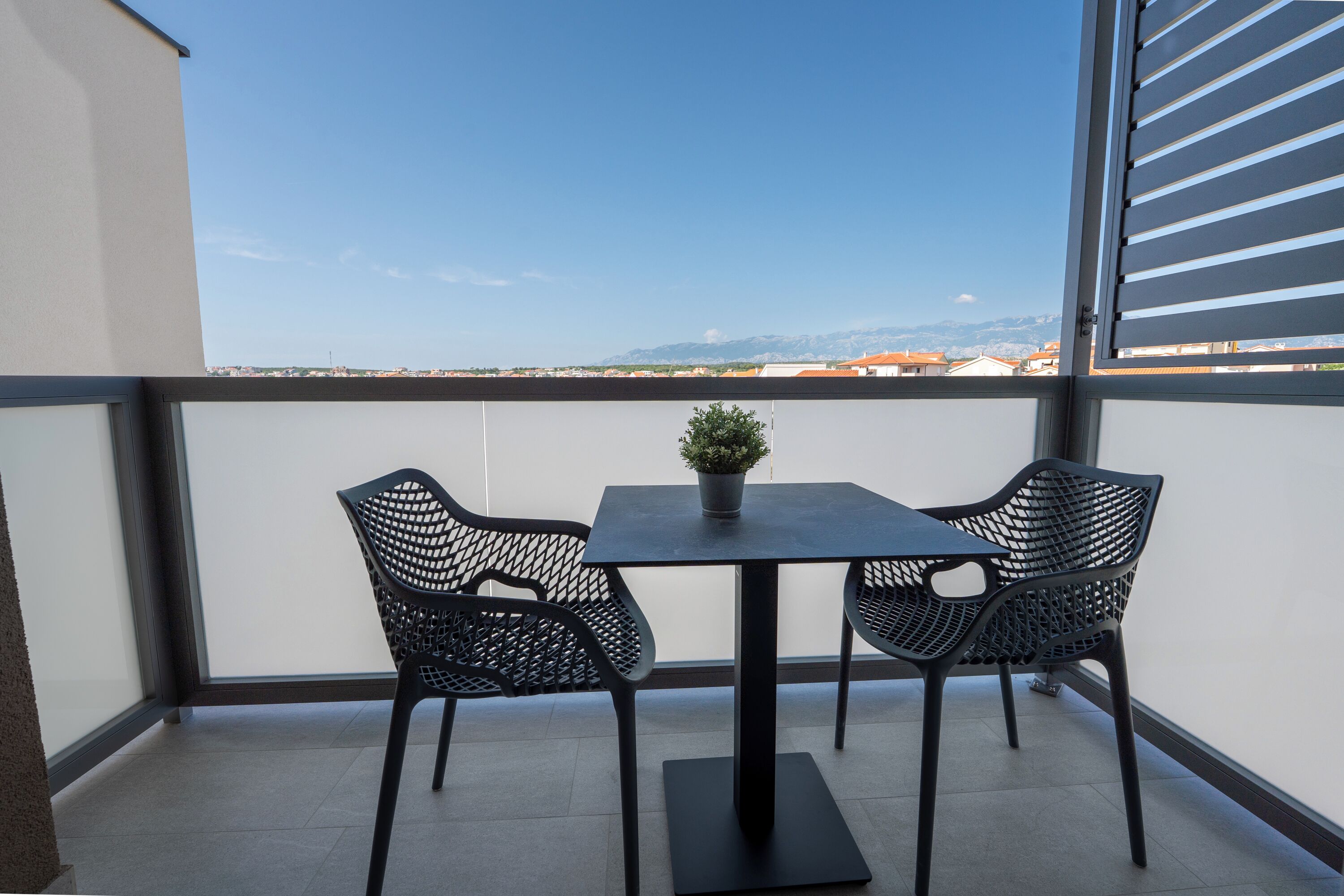 A balcony with a table and two chairs, a potted plant, and a view of distant buildings and mountains.