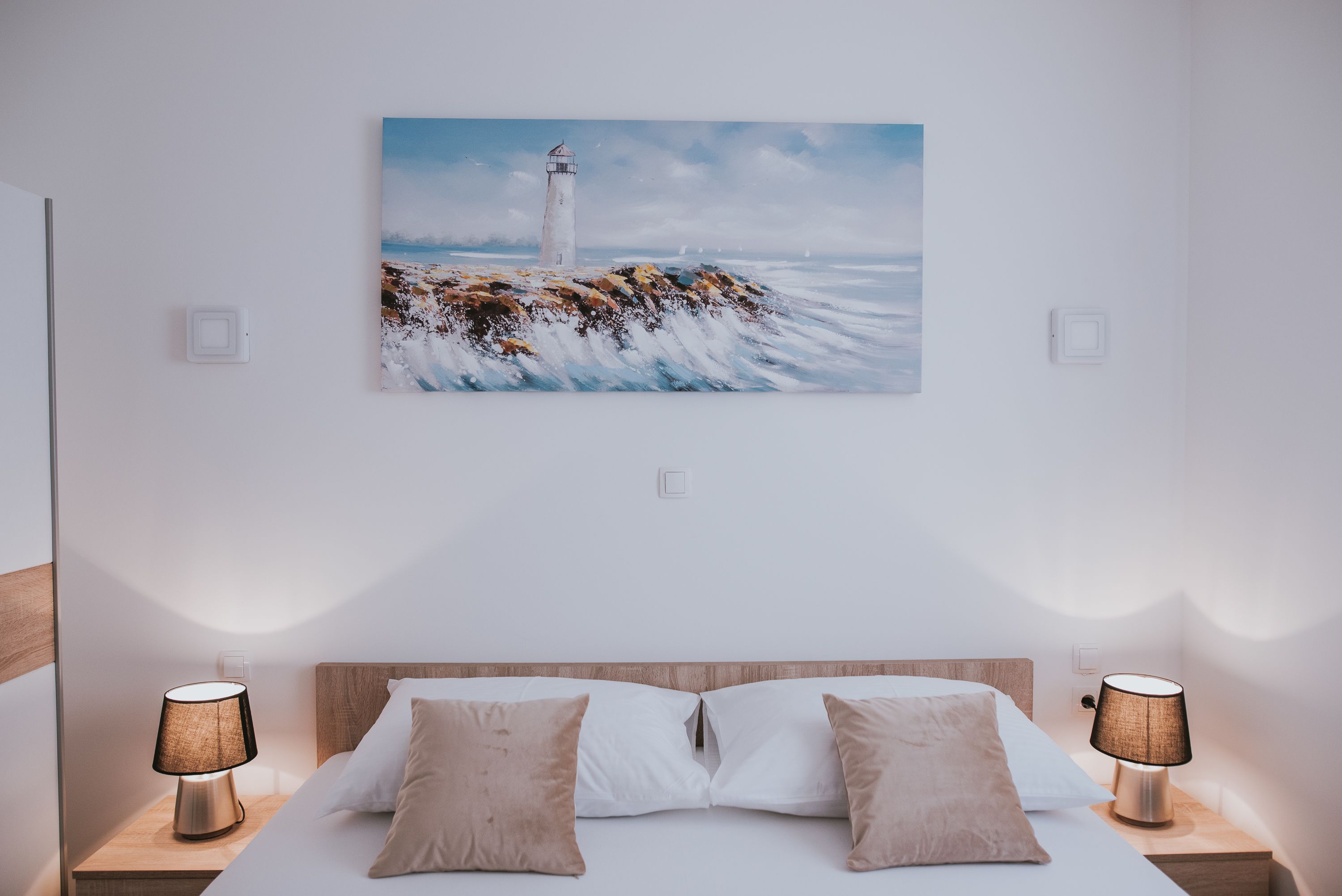 A bedroom with a large bed, two bedside lamps, and a painting of a lighthouse on the wall.