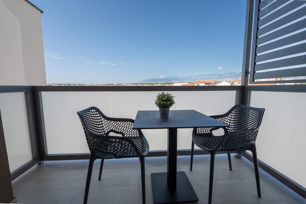 A balcony with a table and two chairs, a potted plant, and a view of distant buildings and mountains.