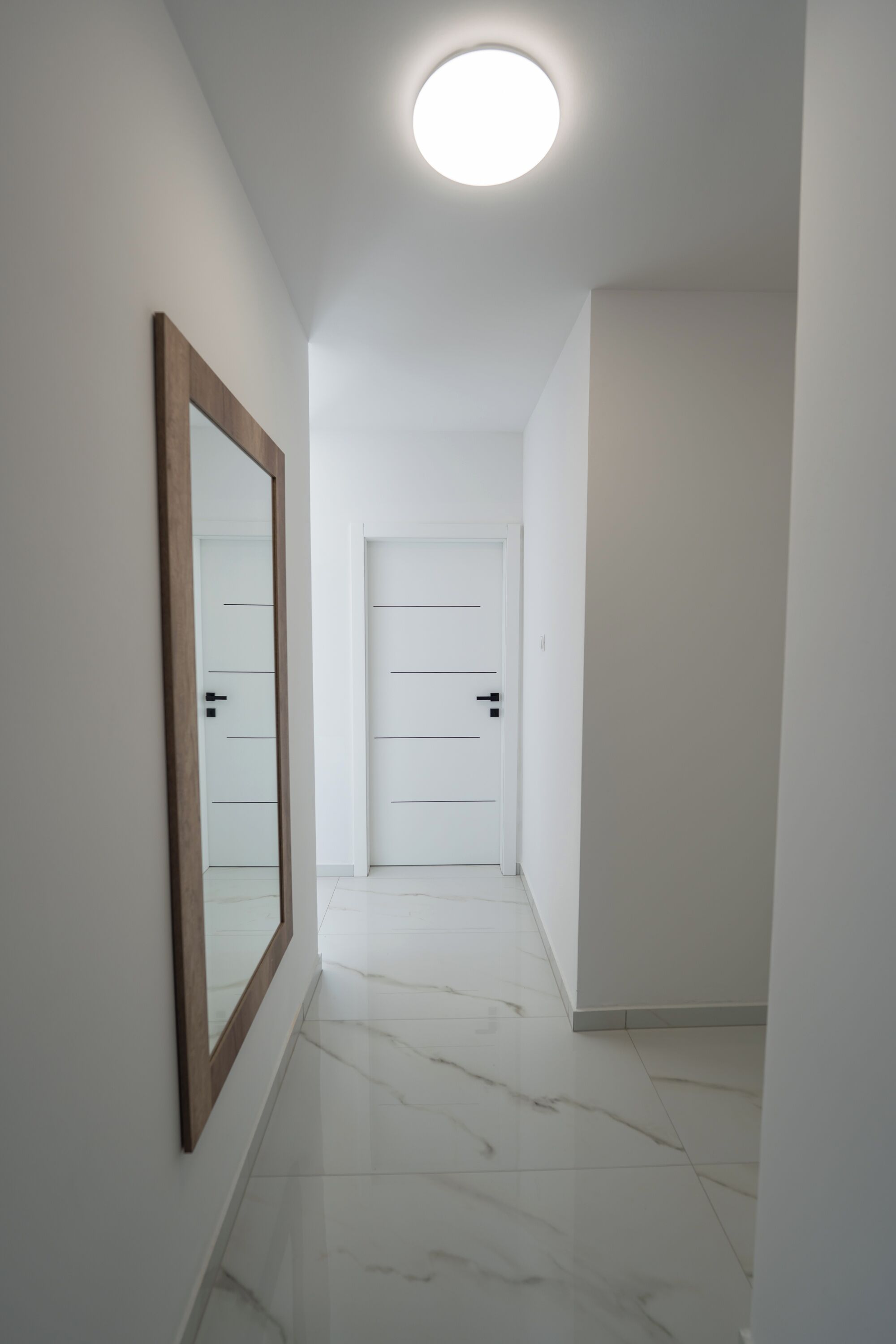A hallway with white walls, a marble floor, and a large mirror on the left.