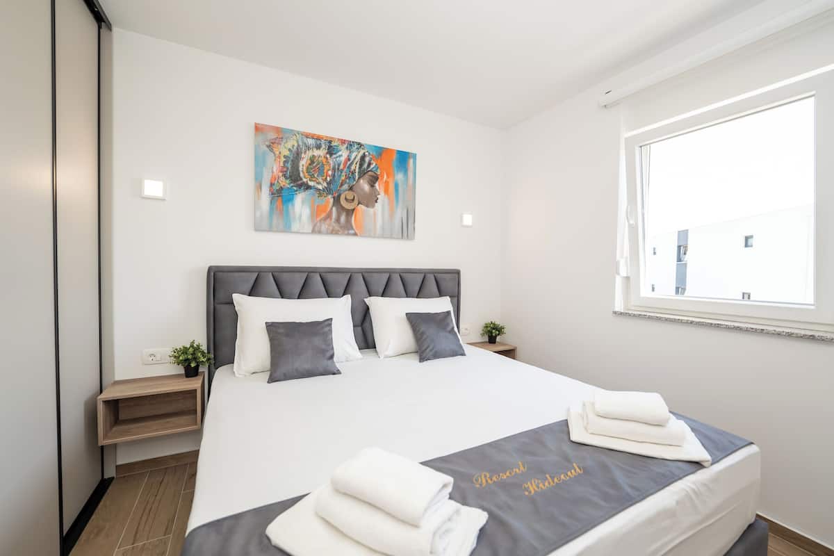 Une chambre d’hôtel moderne avec un grand lit, une tête de lit, un tableau, une fenêtre et une table de chevet agrémentée d’une plante.