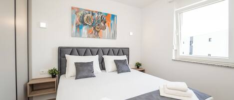 A modern hotel room with a large bed, a headboard, a painting, a window, and a bedside table with a plant.