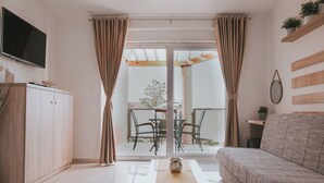 A modern living room with a sofa, a TV mounted on the wall, and a sliding glass door leading to an outdoor area with a table and chairs. - Poolside Hideout Apartments (Novalja)