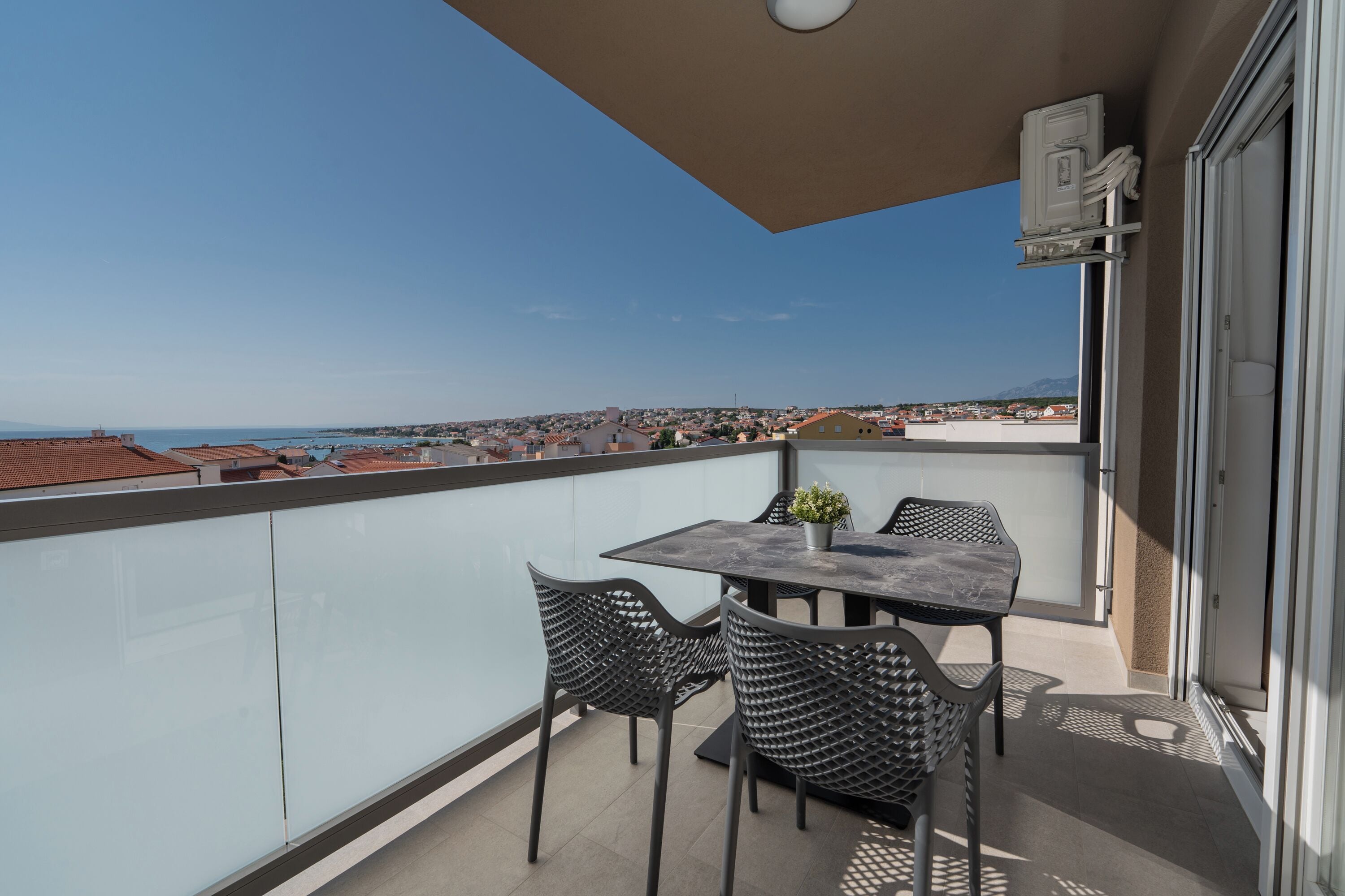 Een balkon met een tafel en stoelen, een bloempot en uitzicht op de zee en gebouwen.