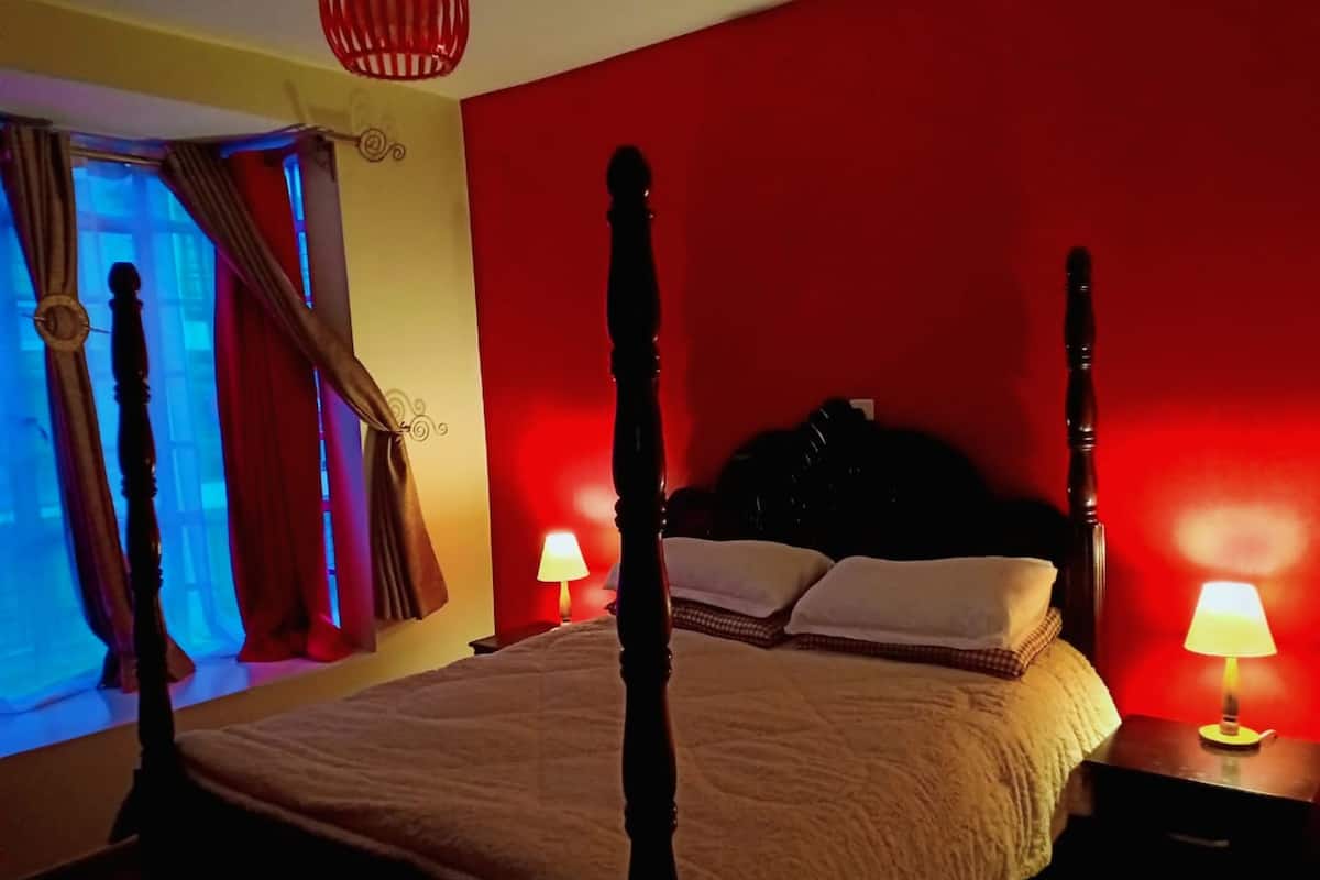 Una cama con dosel, un edredón blanco, dos lámparas de noche y una ventana con cortinas.