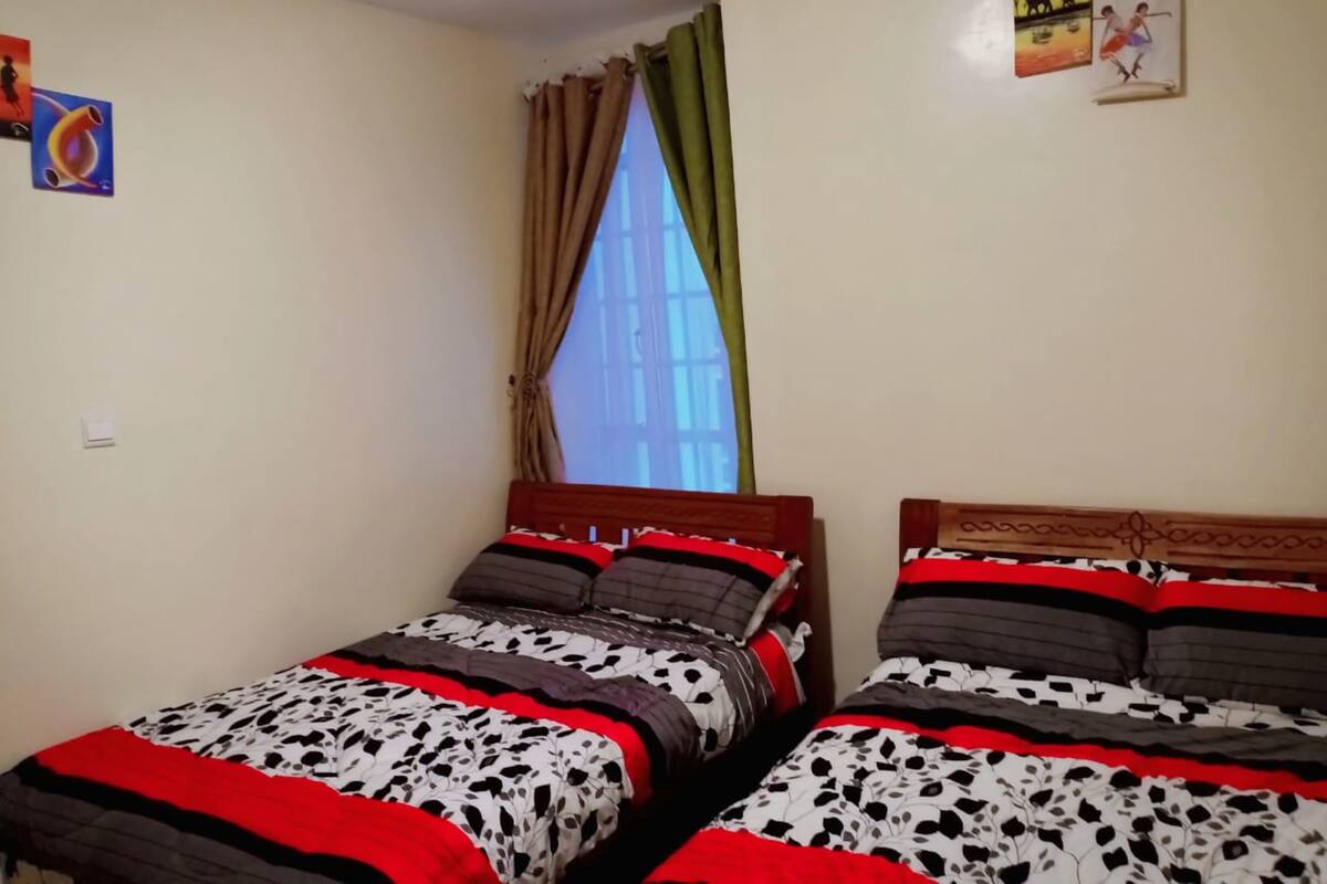 Una habitación con dos camas individuales, cada una con una colcha y almohadas estampadas. Se ve una ventana con cortinas, y obras de arte en las paredes.