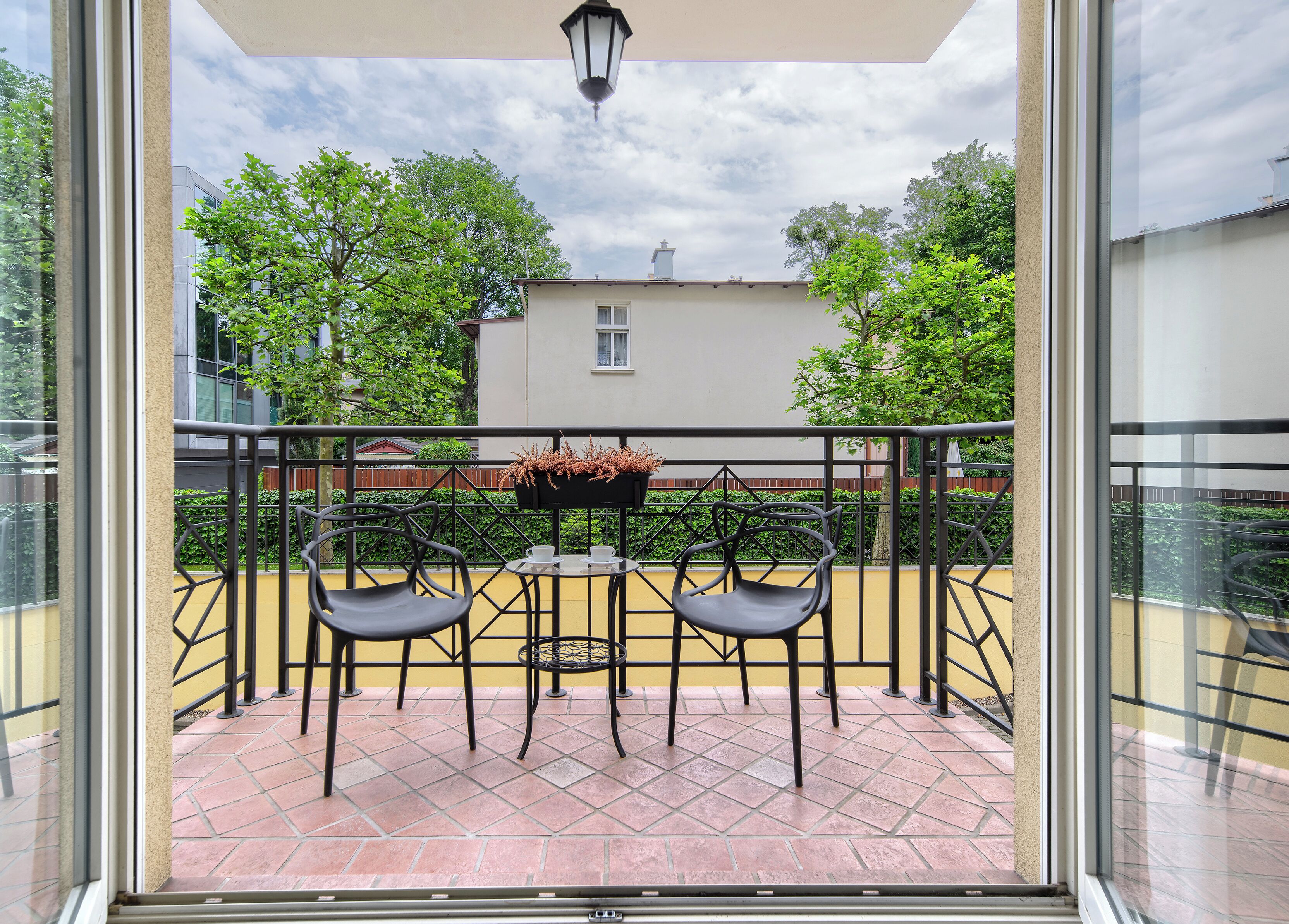 Ein Balkon mit zwei Stühlen und einem Tisch, einem hängenden Blumentopf und Blick auf Bäume und Gebäude.