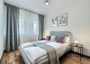 A bedroom with a bed, a bedside table with a lamp, a window with curtains, and two wall art pieces. - Imperial Apartments - Patio Mare (Sopot)