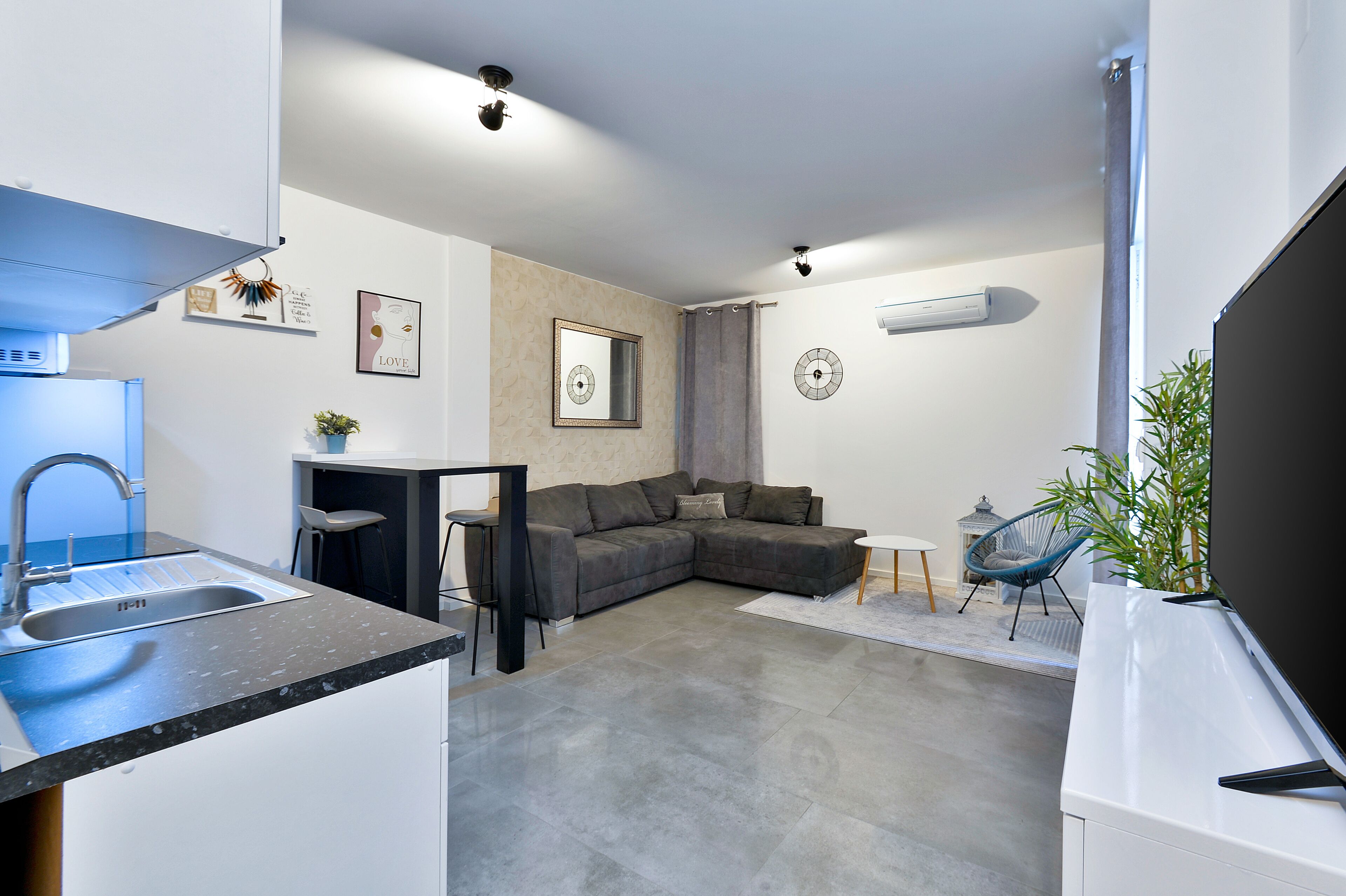 Un espacio habitable compacto con kitchenette, un sofá gris, un pequeño comedor y un televisor.