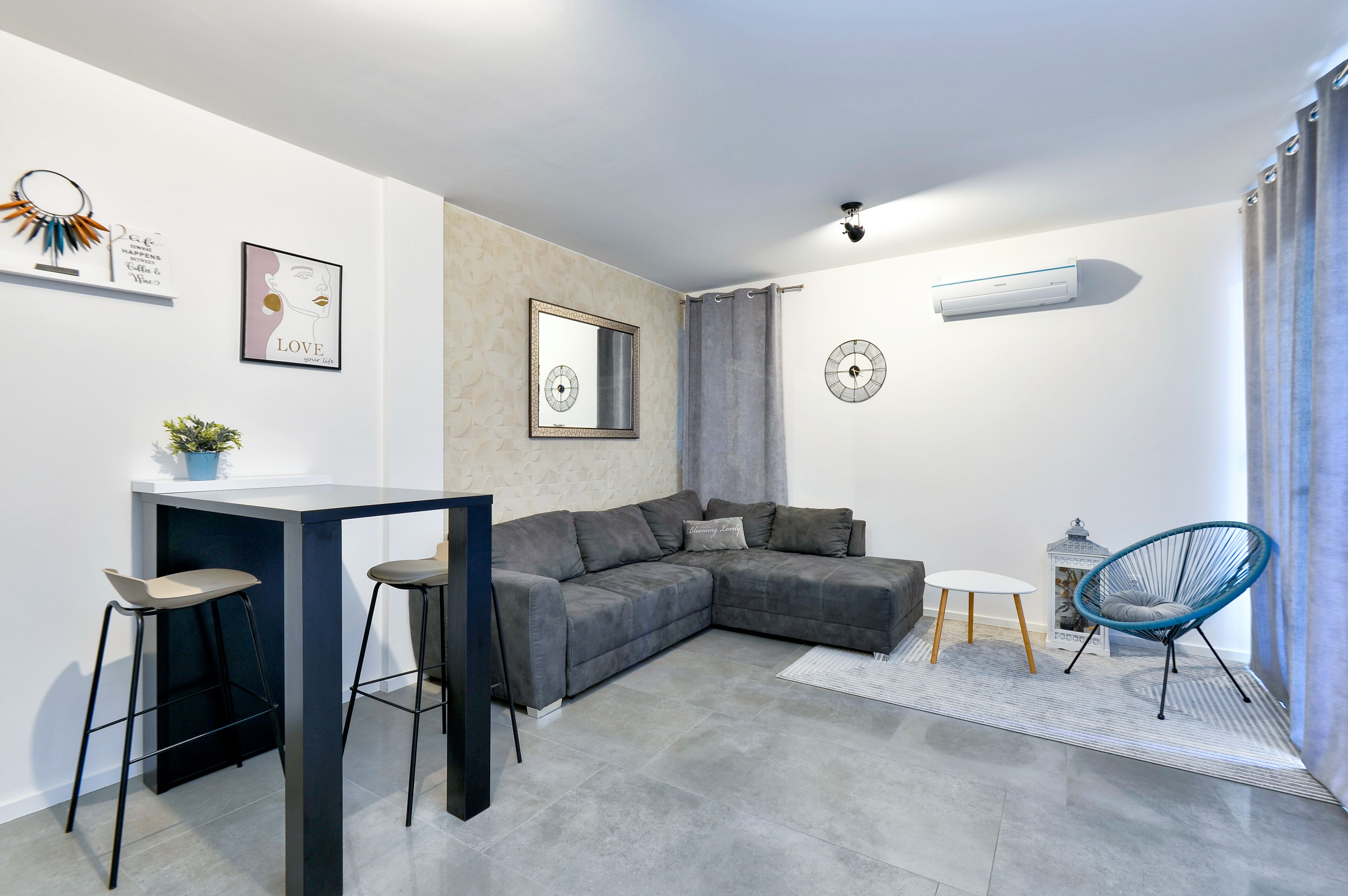 A modern living room with a grey sectional sofa, a small white table, two bar stools, a blue chair, and a decorative mirror.