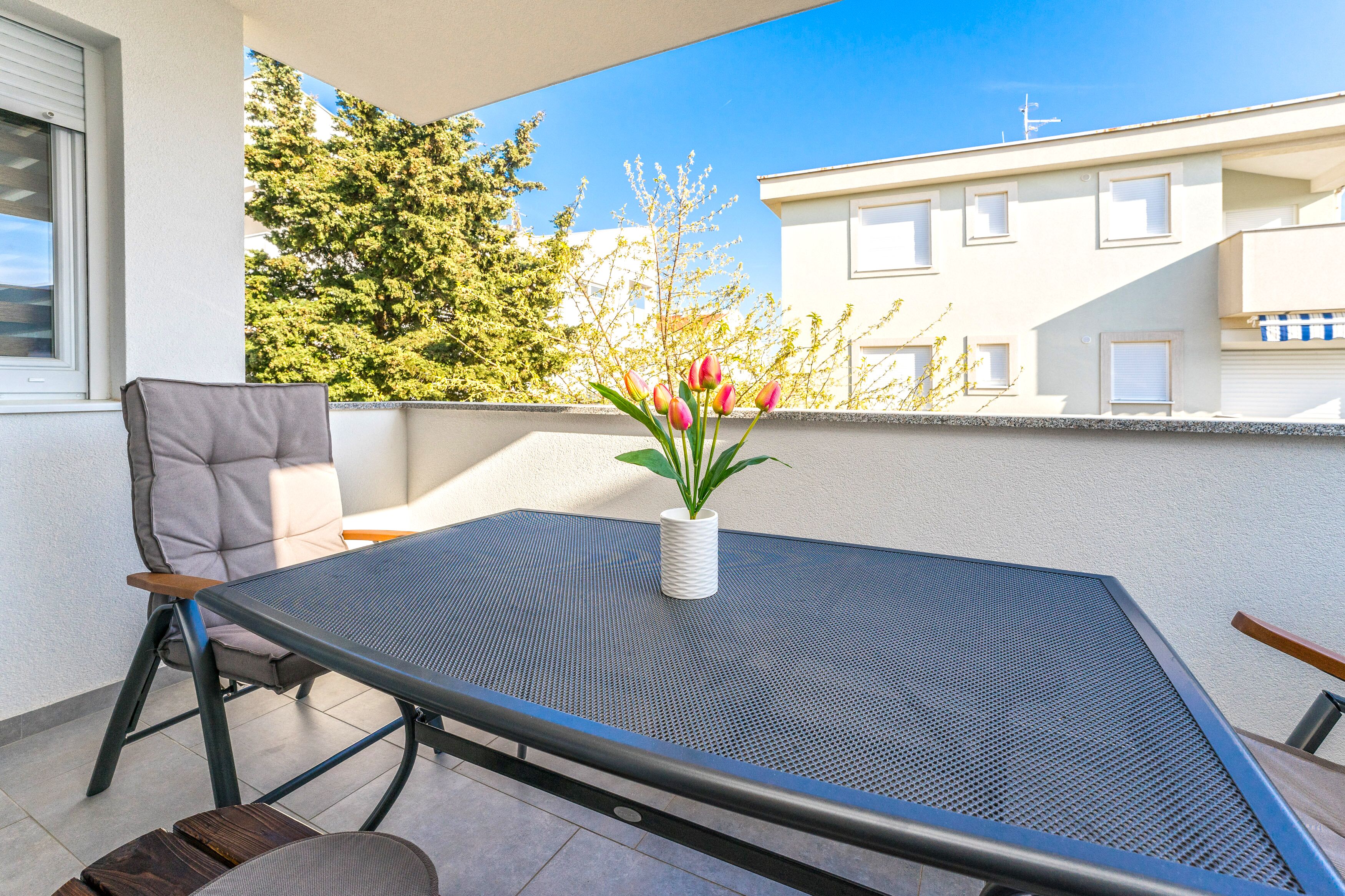 A balcony with a table and chairs, a potted plant, and a view of buildings and trees.