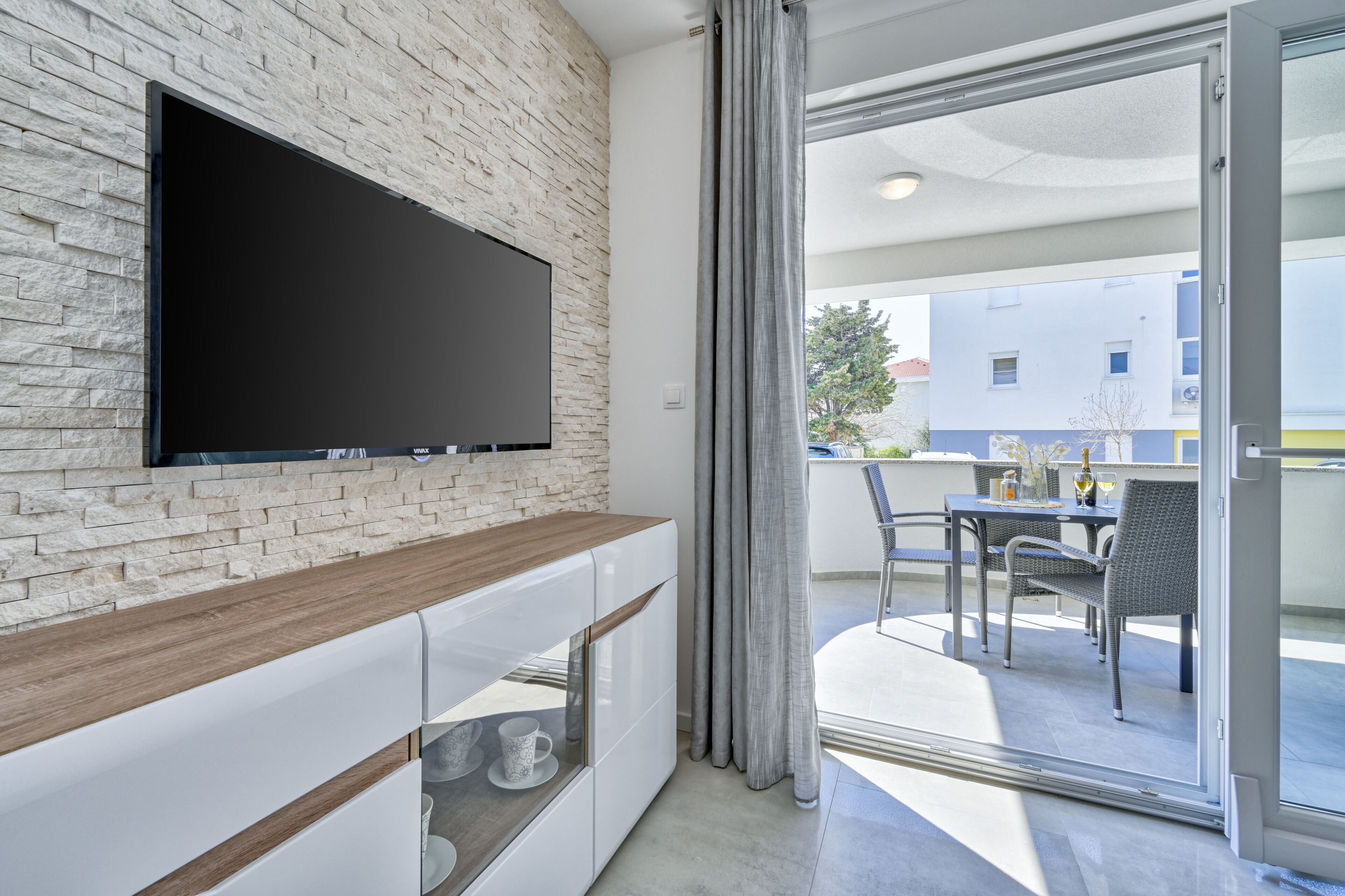 Ein modernes Wohnzimmer mit Flachbildfernseher an einer Ziegelwand, einem Holztisch und einer Schiebetür zu einer Außenpatio-Terrasse mit Esstisch und Stühlen.