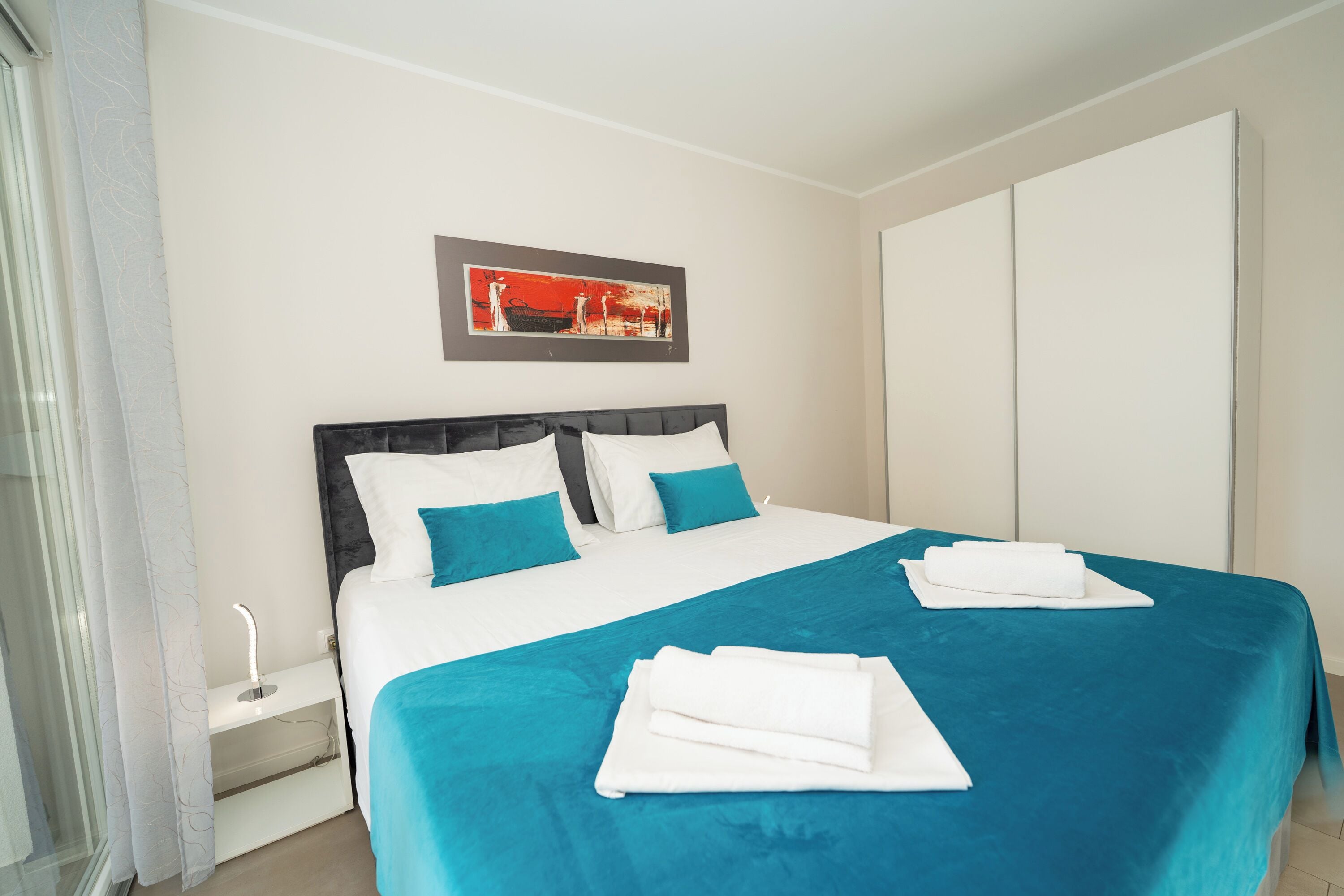 A neatly made bed with white and teal bedding, a bedside table with a lamp, and a painting on the wall.