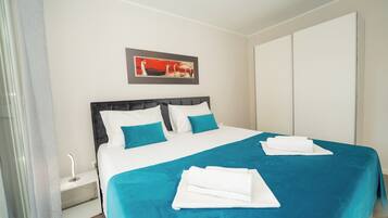A neatly made bed with white and teal bedding, a bedside table with a lamp, and a painting on the wall.