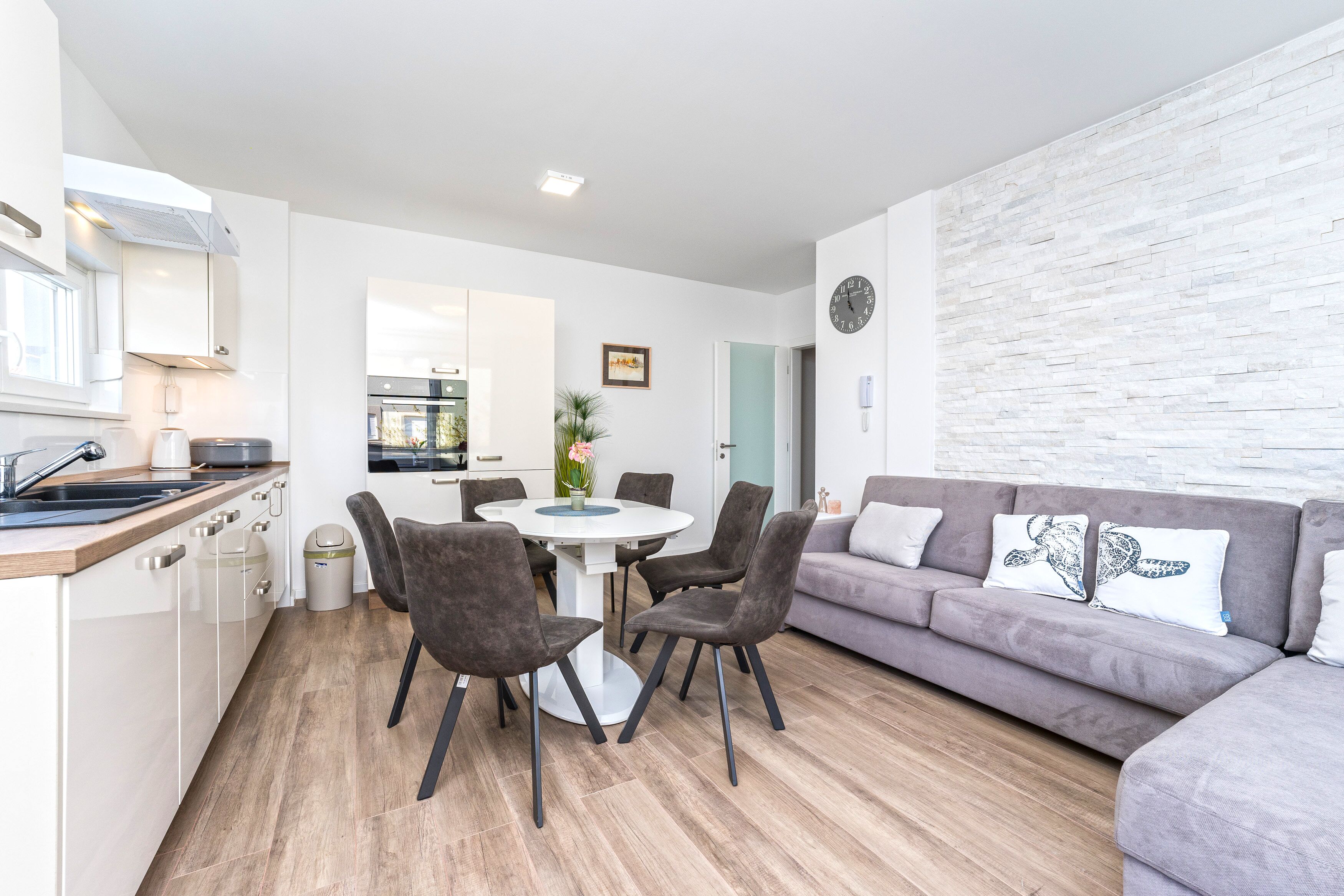 A modern kitchen with white cabinets, a dining table with chairs, and a grey sofa with decorative pillows.