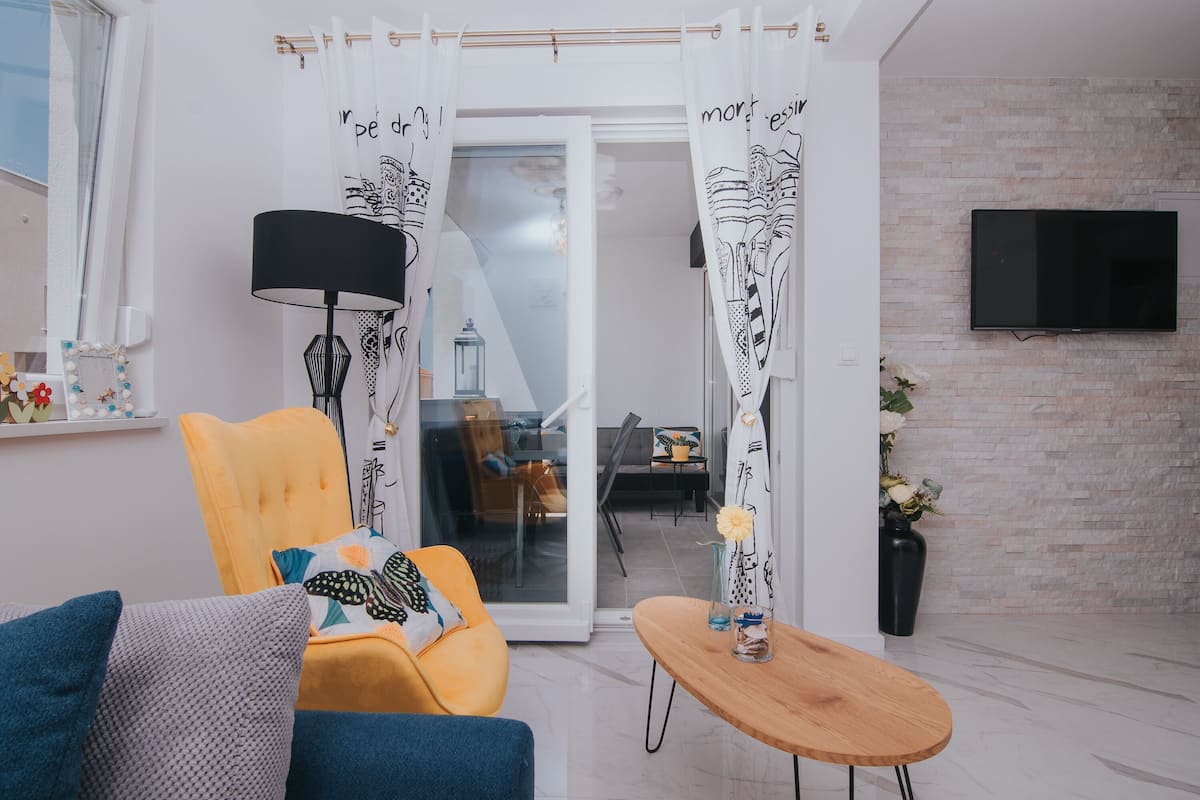 A modern living room with a yellow armchair, a blue sofa, a wooden side table, and a glass door leading to a dining area.