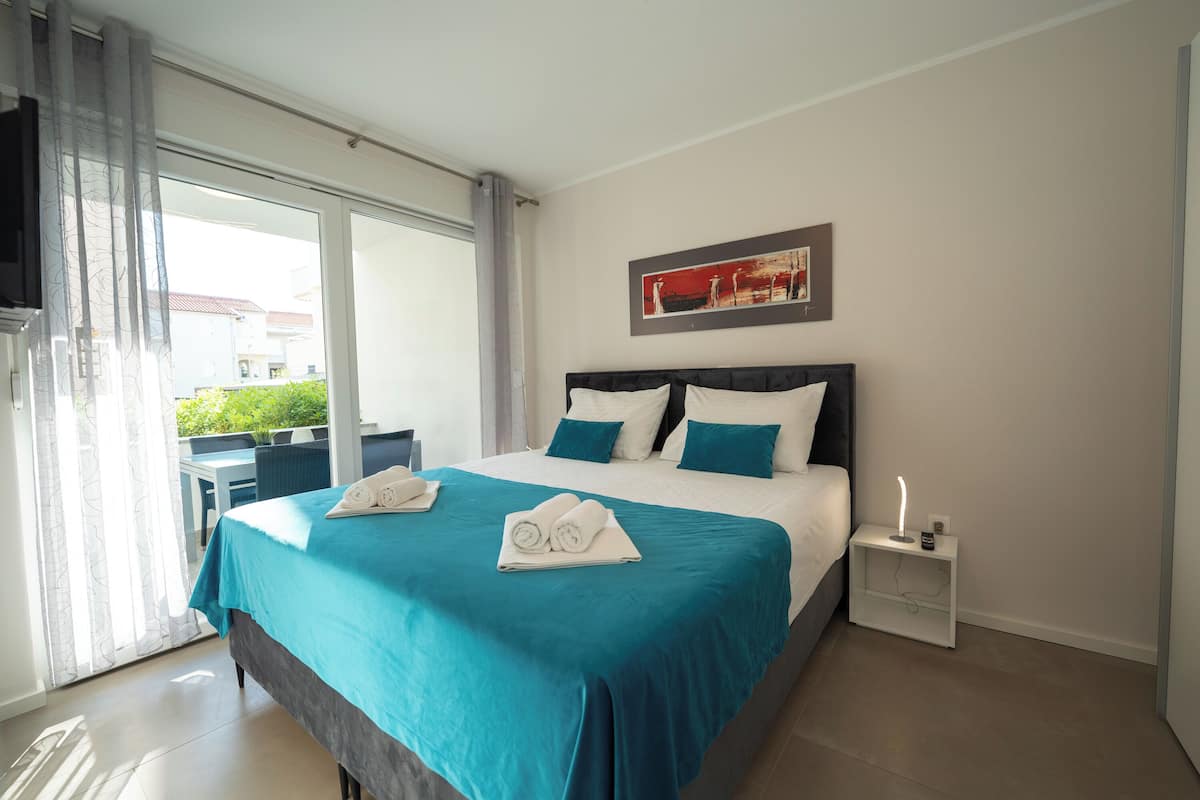 A hotel room with a bed, a nightstand, a TV, and a view of the outside.