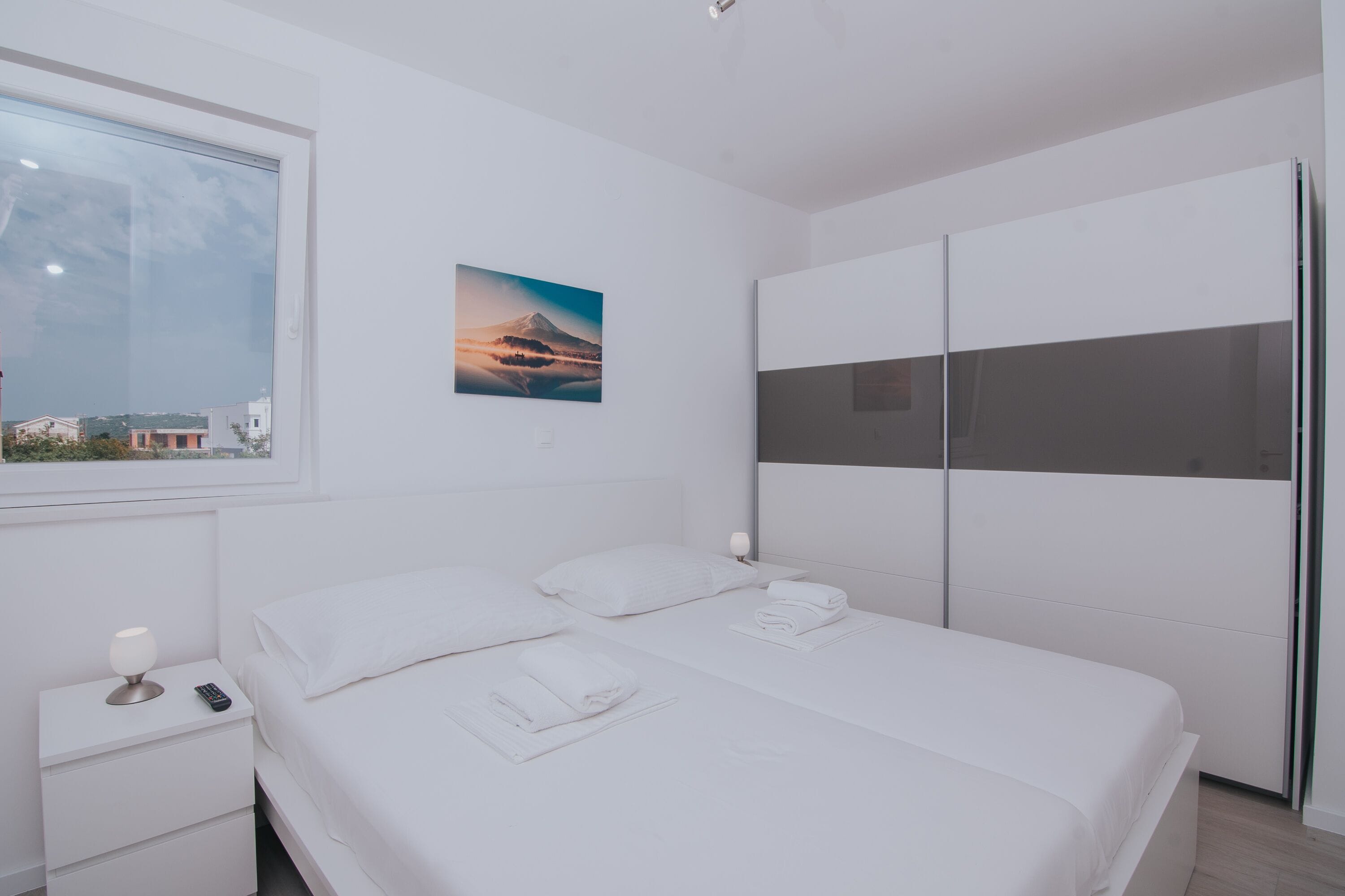 A modern hotel room with a large bed, a bedside table with a lamp, a remote control, neatly folded towels, a painting of a mountain, and a wardrobe.