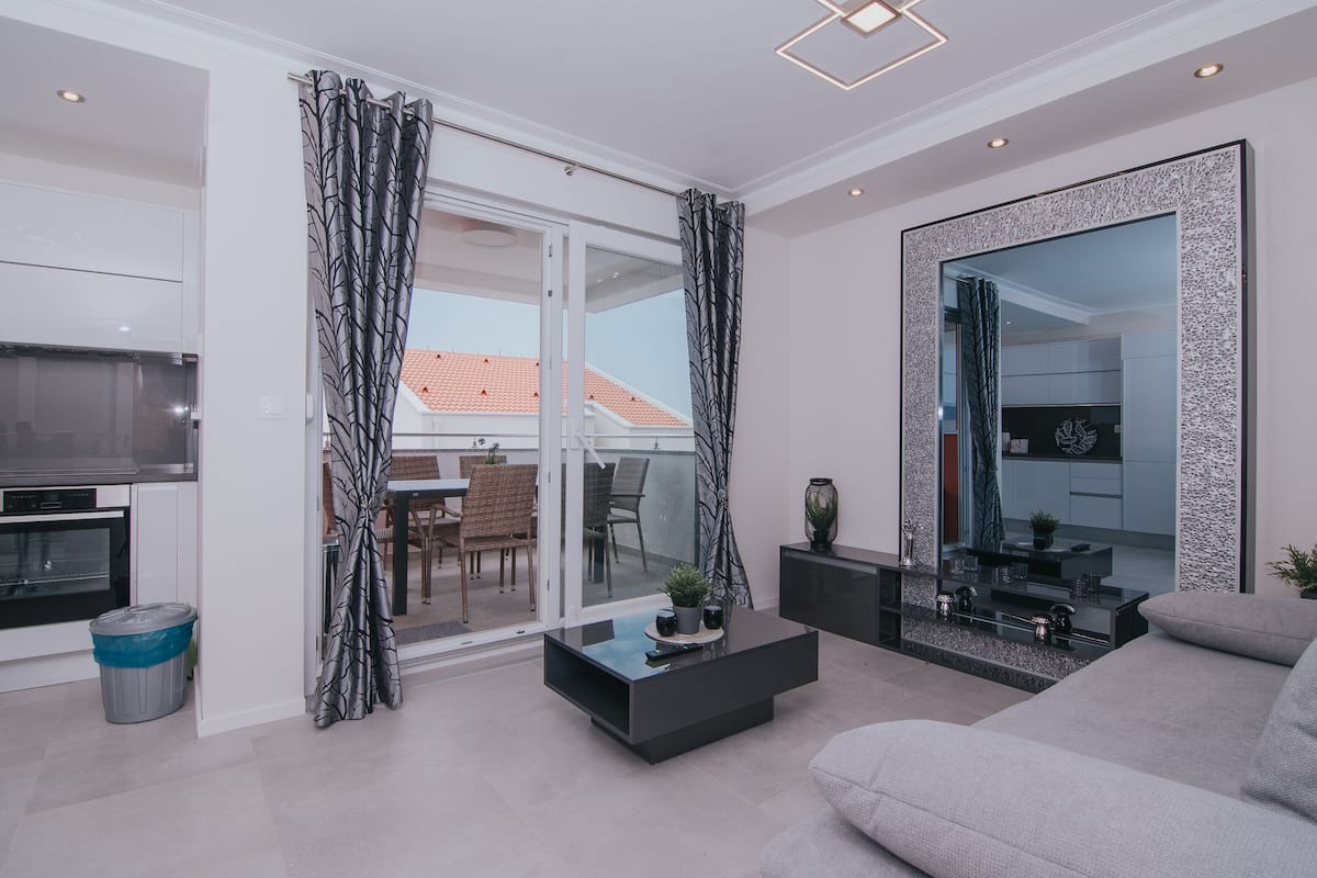 A modern living room with a sofa, a coffee table, and a TV. There is a sliding glass door leading to an outdoor area with a table and chairs.