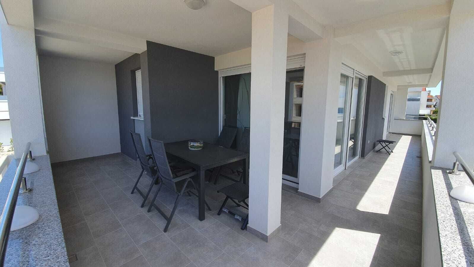A modern outdoor dining area with a table and chairs, a glass door leading to a covered space, and a tiled floor.