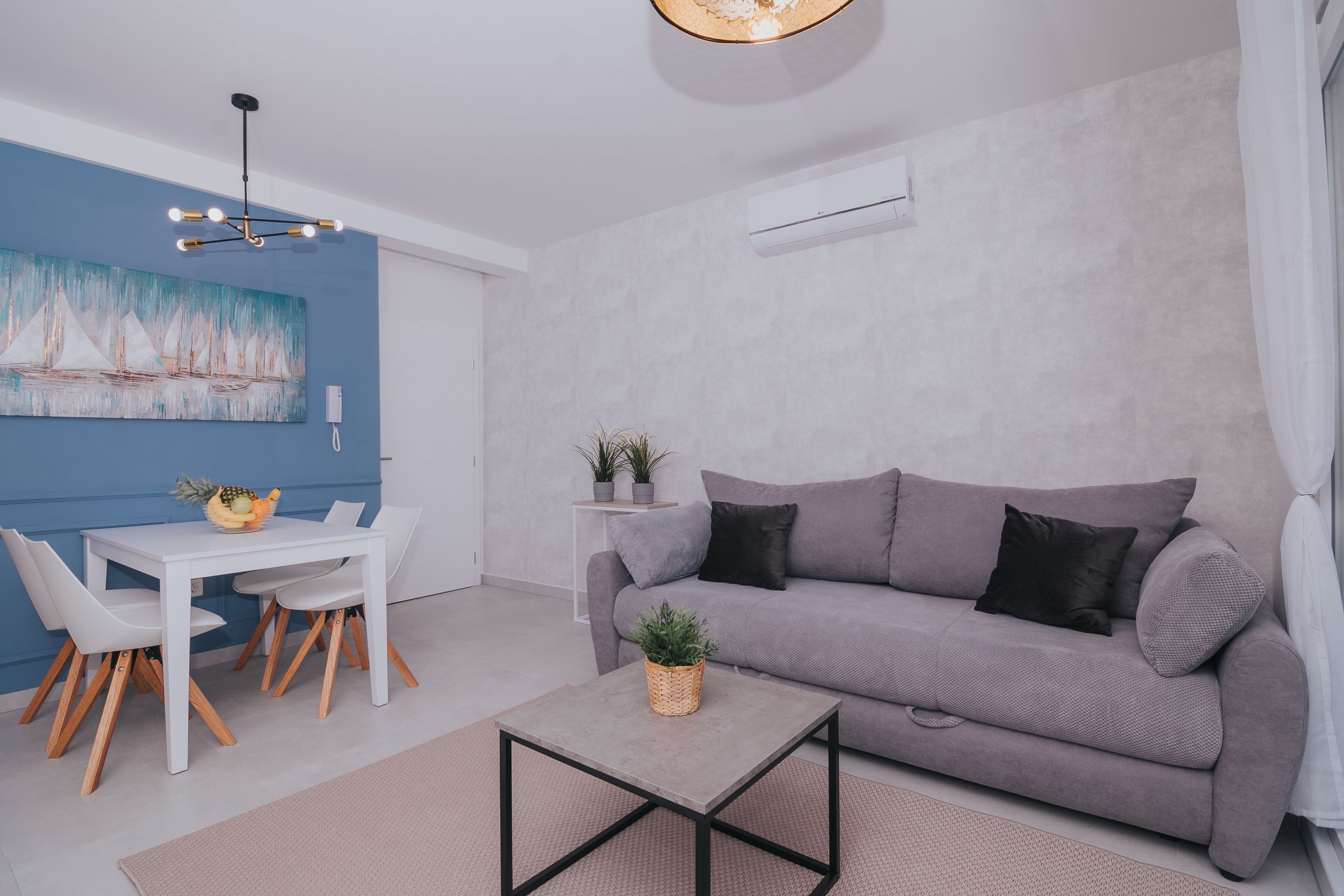 A modern living room with a grey sofa, white dining table, and a ceiling light fixture.