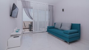 Een moderne woonkamer met een blauwgroene bank, een flatscreen televisie aan de muur en een klein wit bijzettafeltje met een schaal en afstandsbediening.