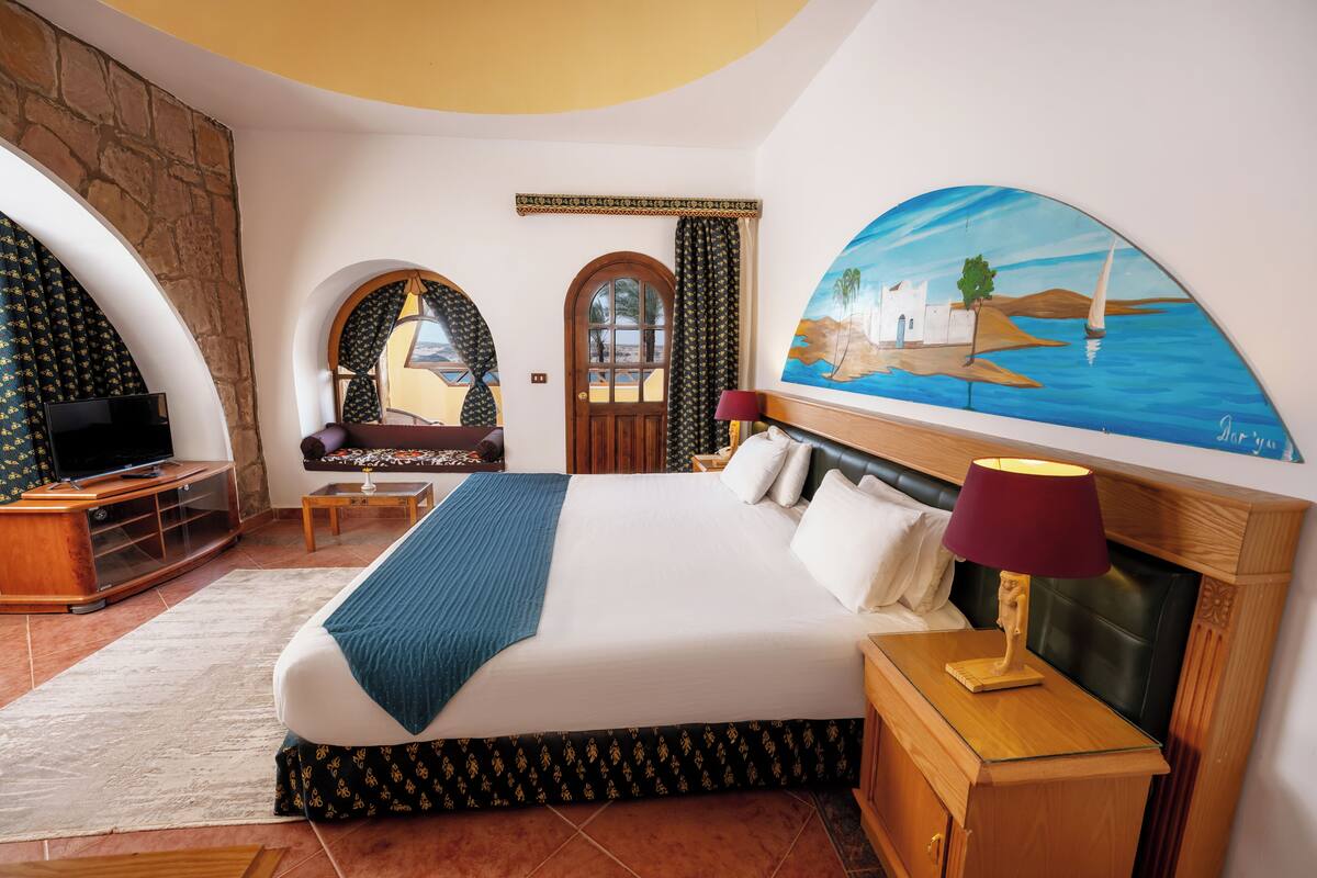 A hotel room with a large bed, a TV, a wooden nightstand, and a painting of a coastal scene.