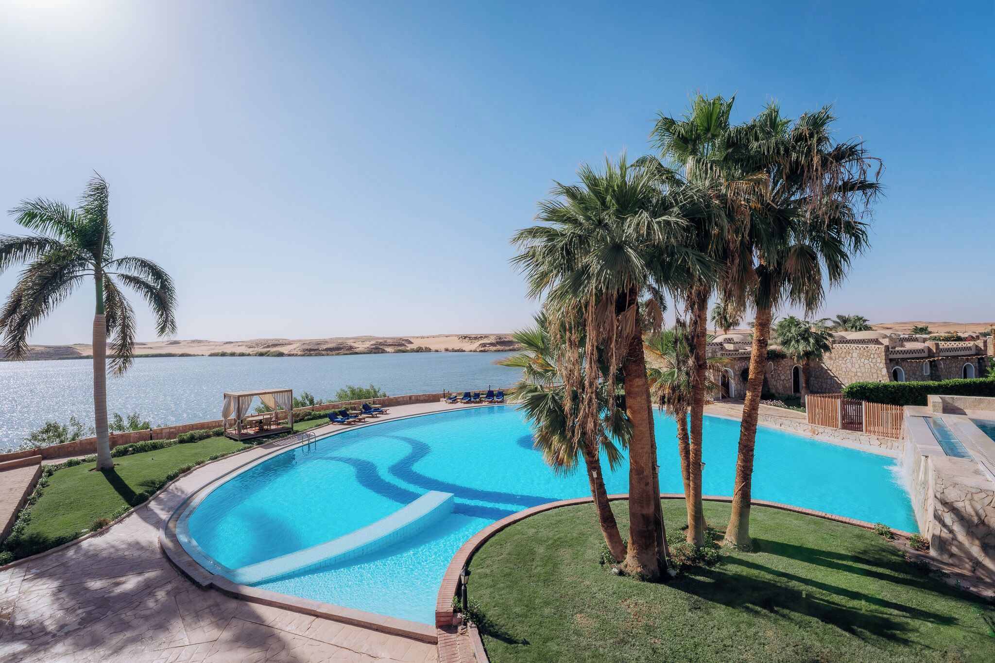 Piscina con vista limpida sull'acqua, circondata da palme e un edificio in pietra.