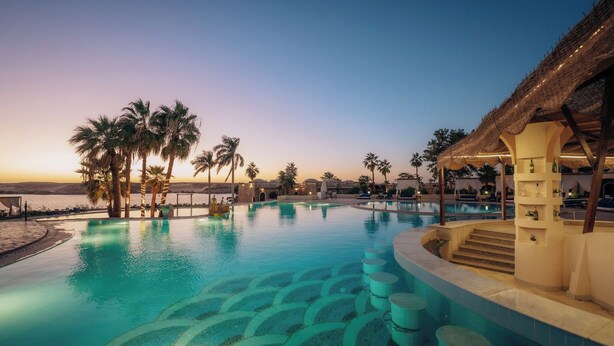 Une piscine avec vue sur le coucher de soleil, des palmiers et une structure surmontée d’un toit de chaume.