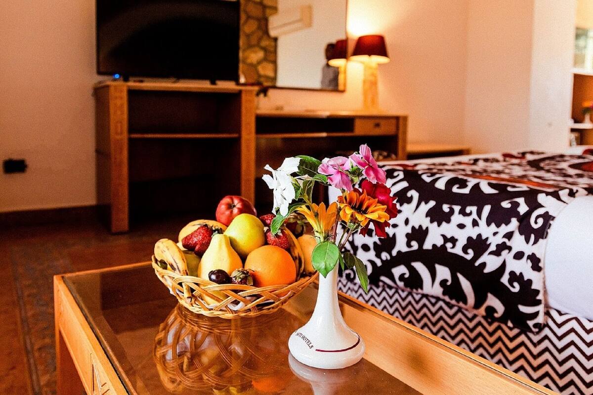 A room with a wooden table, a fruit basket, a vase with flowers, a patterned bedspread, and a fireplace.