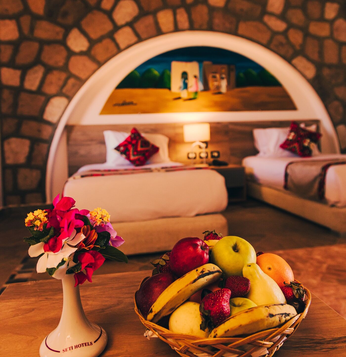 A hotel room with a stone wall, a bed with a headboard, a bedside table with a vase of flowers, and a fruit basket.