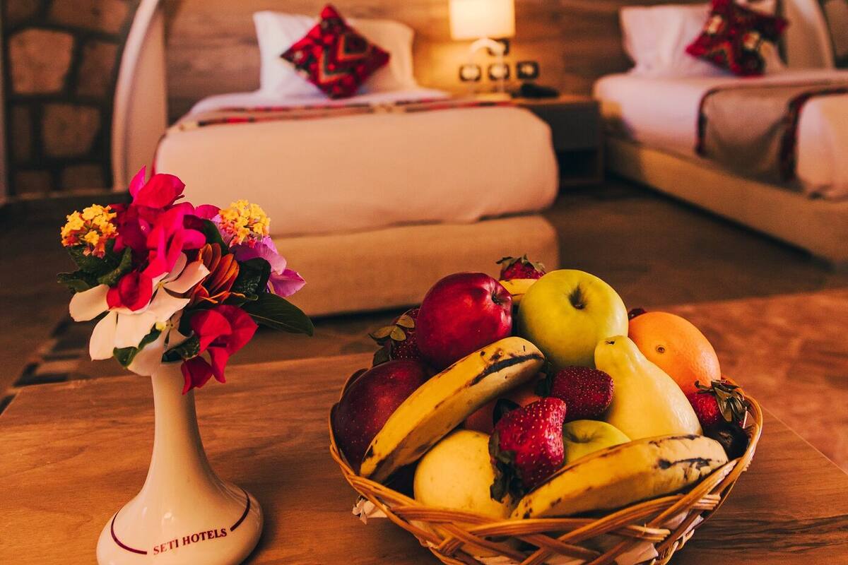 A hotel room with a stone wall, a bed with a headboard, a bedside table with a vase of flowers, and a fruit basket.