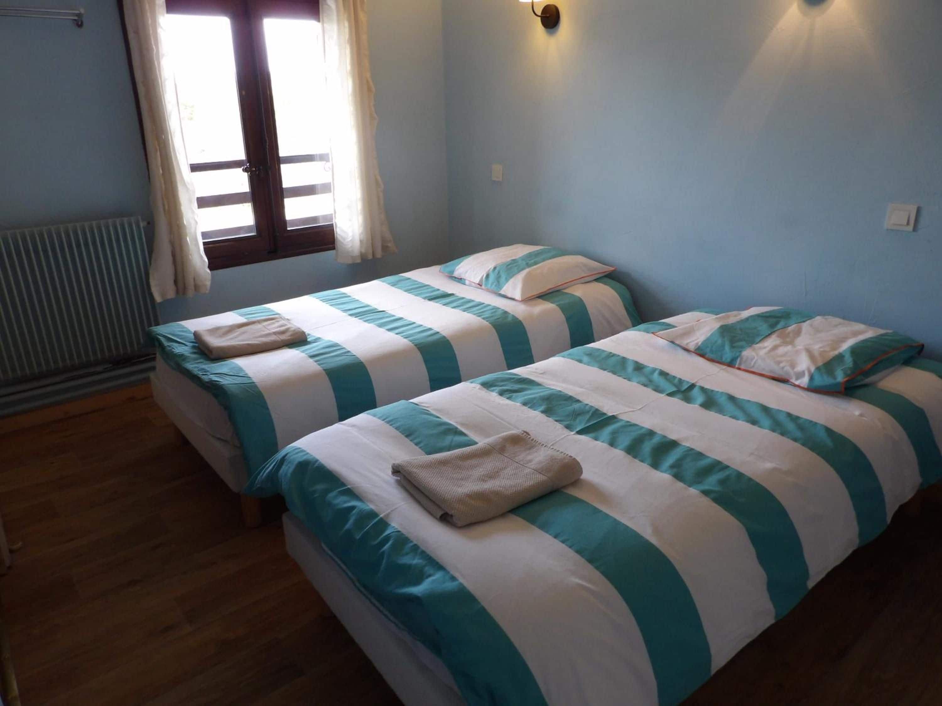 Una habitación con dos camas, cada una con ropa de cama y almohadas a rayas, un radiador, una ventana con cortinas y una lámpara fijada a la pared.