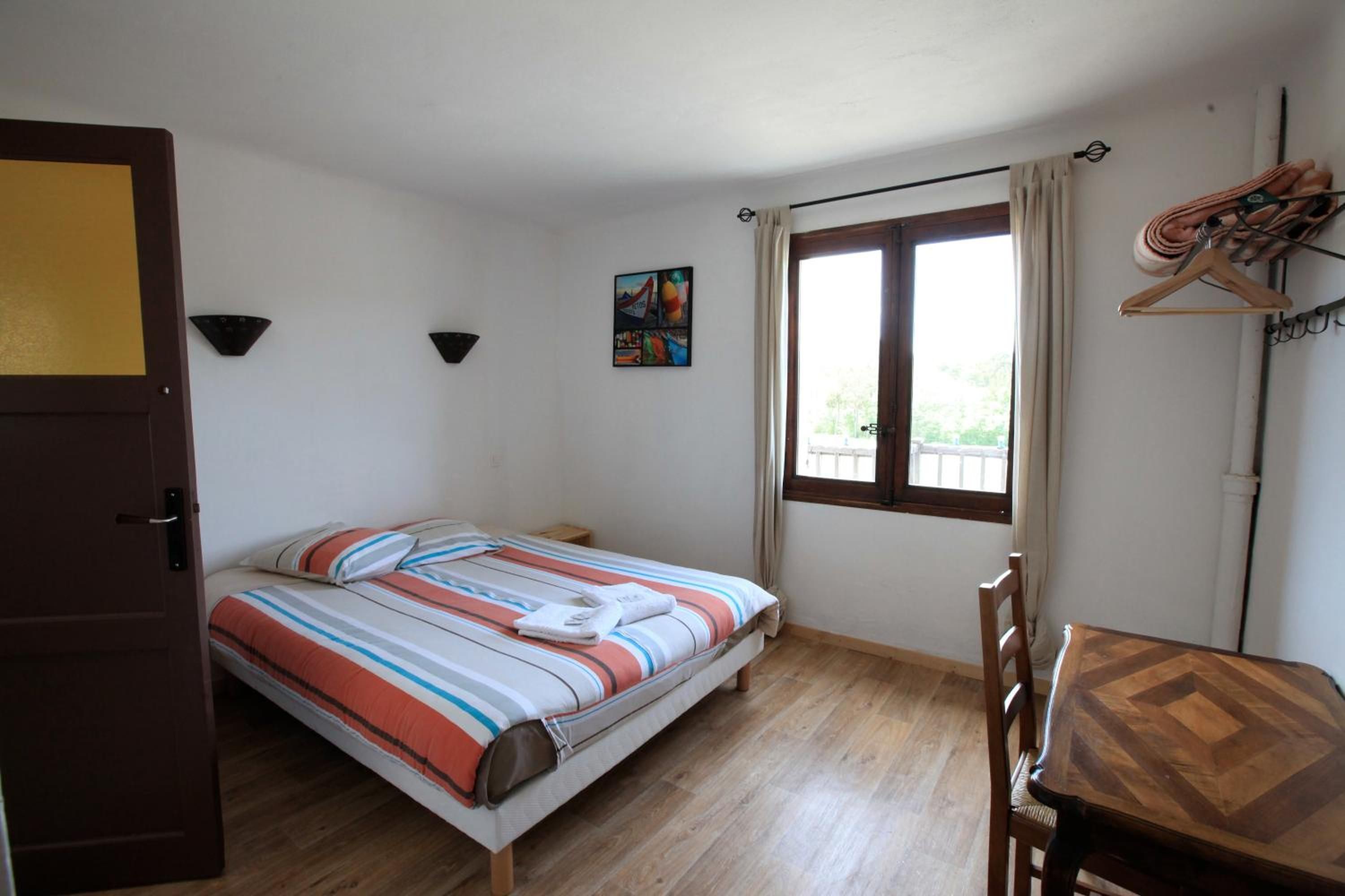 Une chambre avec un lit simple, un plancher en bois, une petite table, une chaise, une fenêtre avec des rideaux et un tableau au mur.