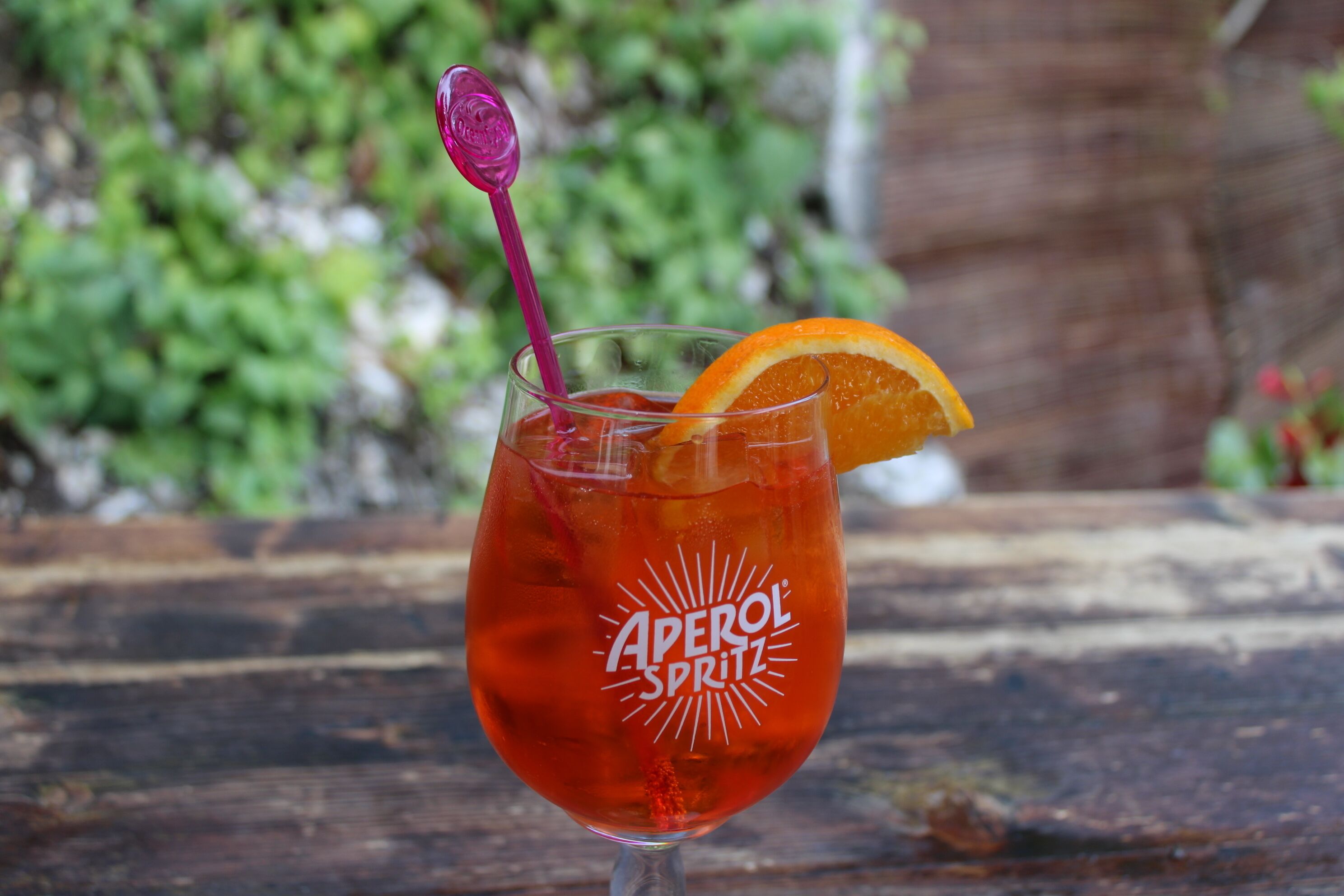Un vaso de Aperol Spritz con una rodaja de naranja y una pajita morada sobre una superficie de madera.